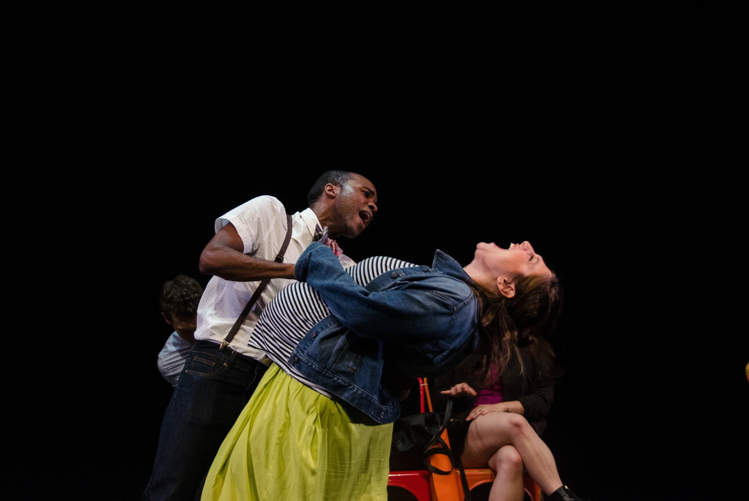 A theatrical stage scene depicting a man in a white shirt and suspenders holding a woman in a striped shirt and a yellow skirt. The man appears to be shouting or singing passionately, and the woman is leaning back with her eyes closed. Other performe