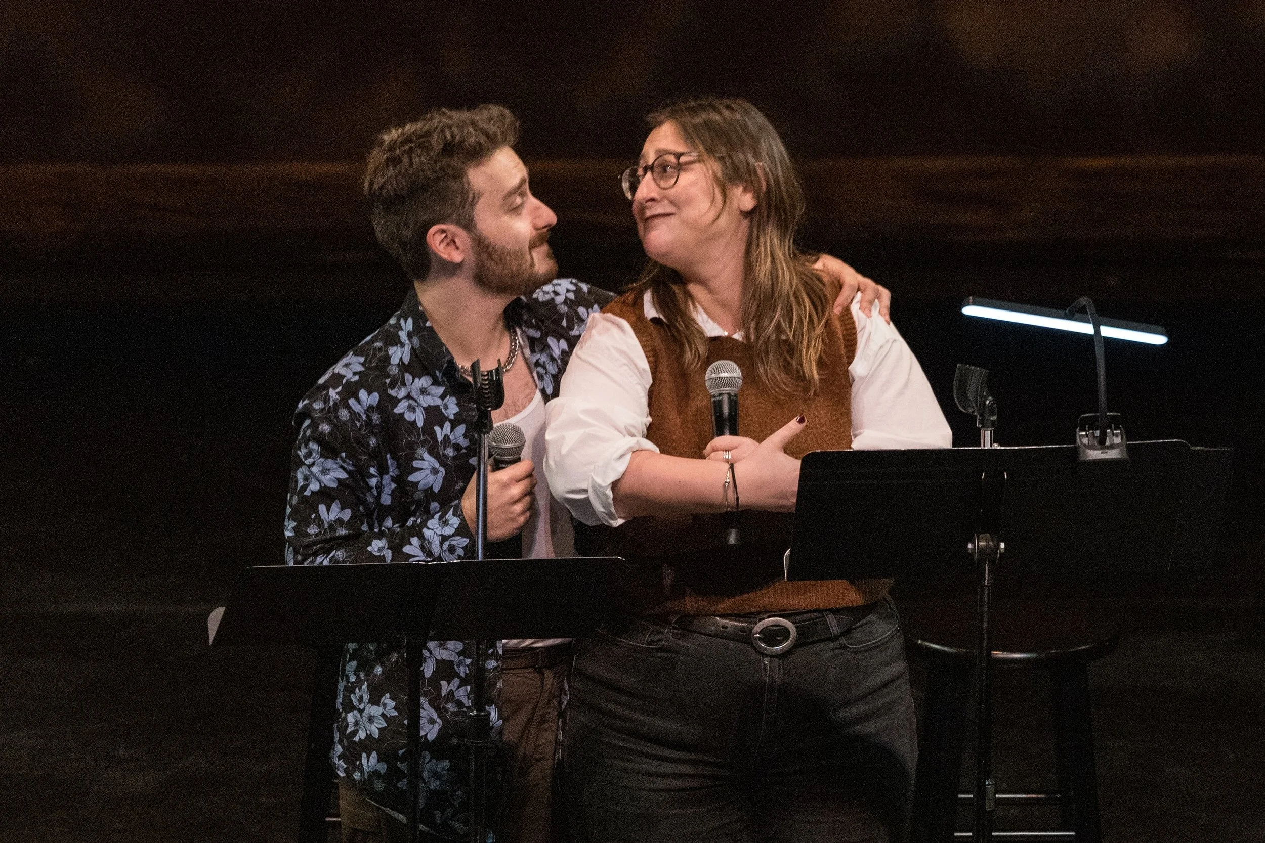 A man and a woman on stage holding microphones and looking at each other, with music stands in front of them, against a dark background.