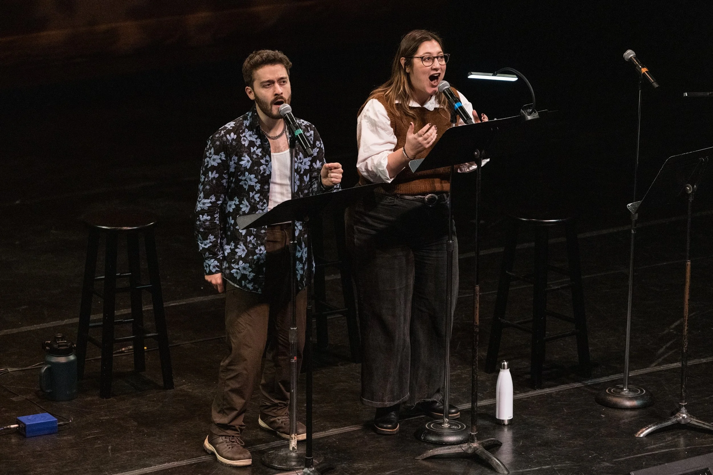 Two people performing on stage, singing into microphones, with music stands and stage lights in the background.