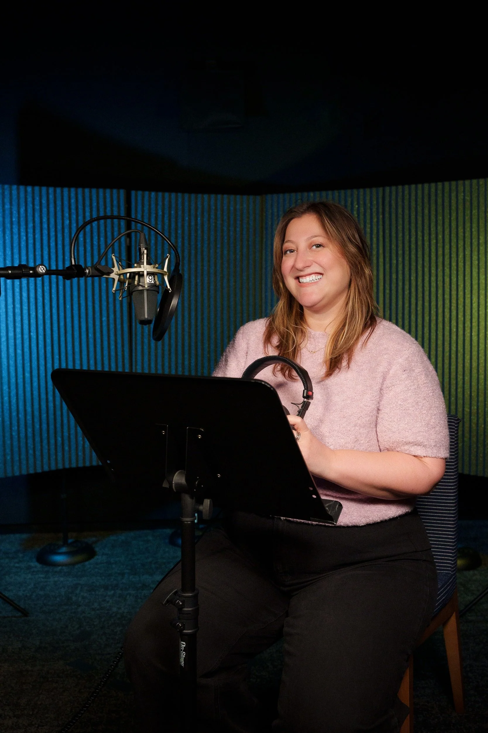 A woman with shoulder-length brown hair, wearing a pink sweater, is smiling while sitting in a recording studio. She is holding a pair of headphones and is positioned in front of a music stand and a professional microphone on a stand. The background features green and blue acoustic panels.