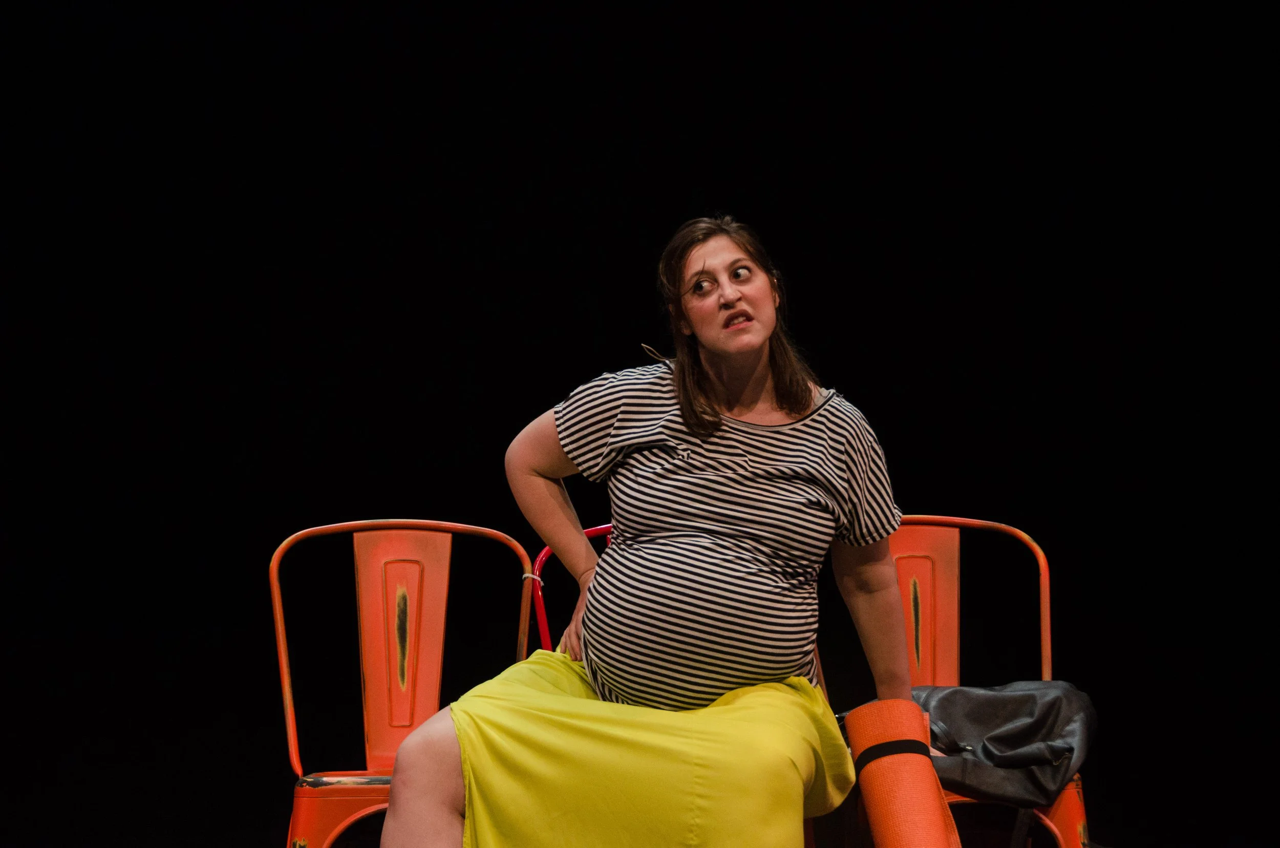 A woman with brown hair, wearing a black and white striped shirt and yellow pants, sitting on a chair on a black background, looking to her left with an expressive face.