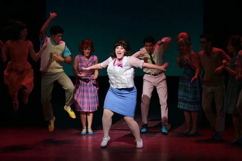 Stage performance with actors in colorful costumes, woman in center dancing and smiling, others around her in playful poses.