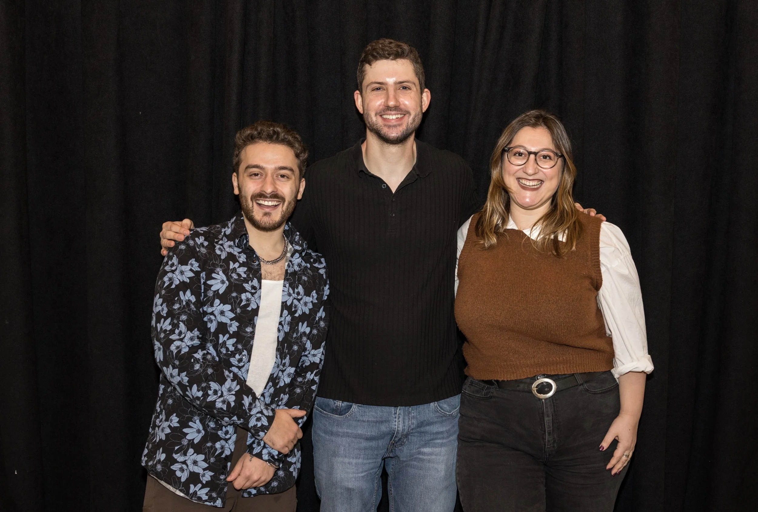 Three people smiling, standing in front of a black curtain. The person on the left has short curly hair, a beard, and is wearing a patterned shirt. The person in the middle has short dark hair, a beard, and is wearing a black shirt. The person on the