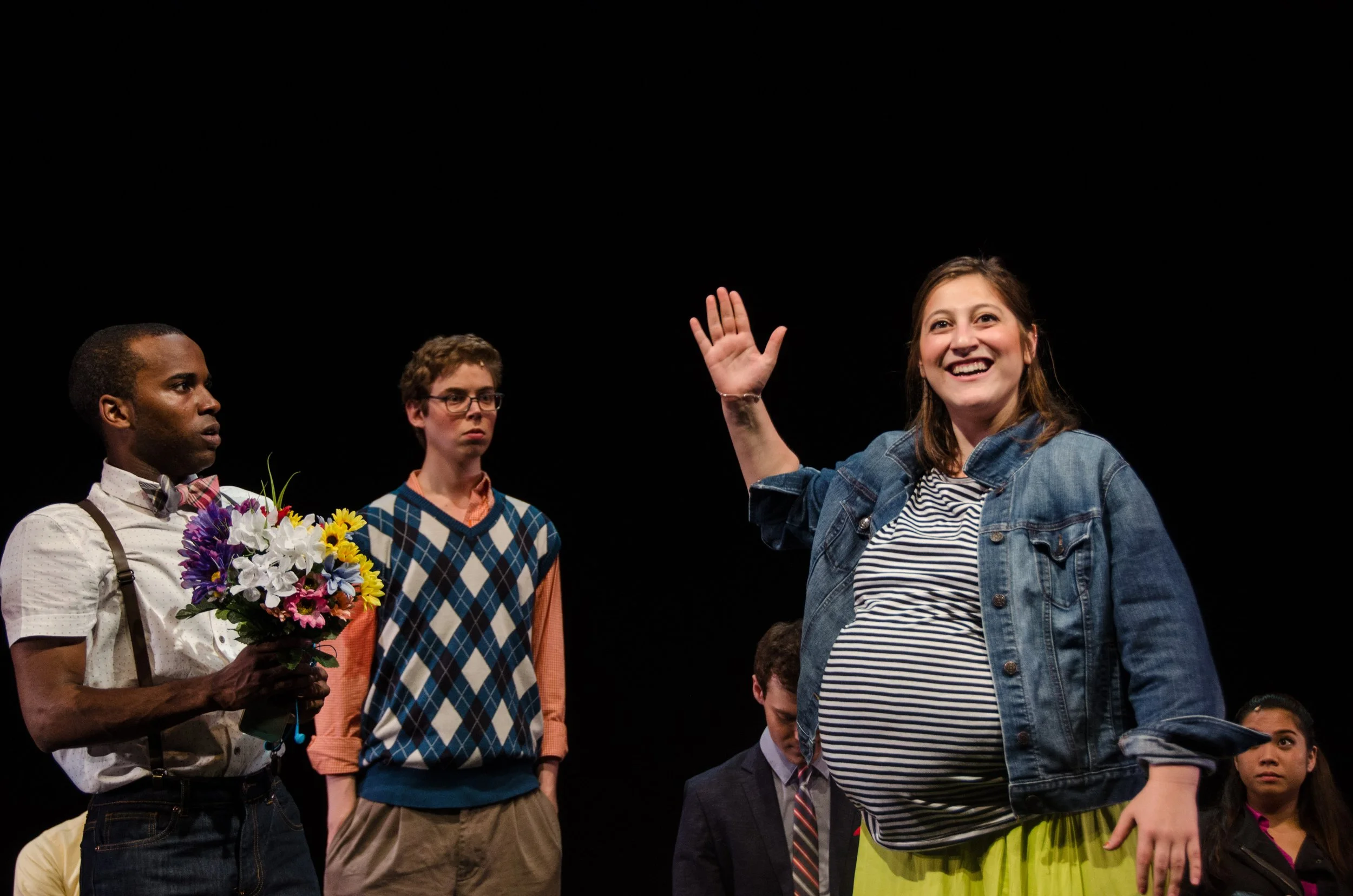 A group of diverse people on stage, with a woman smiling and waving, holding a microphone, and others standing around her, some with serious expressions.