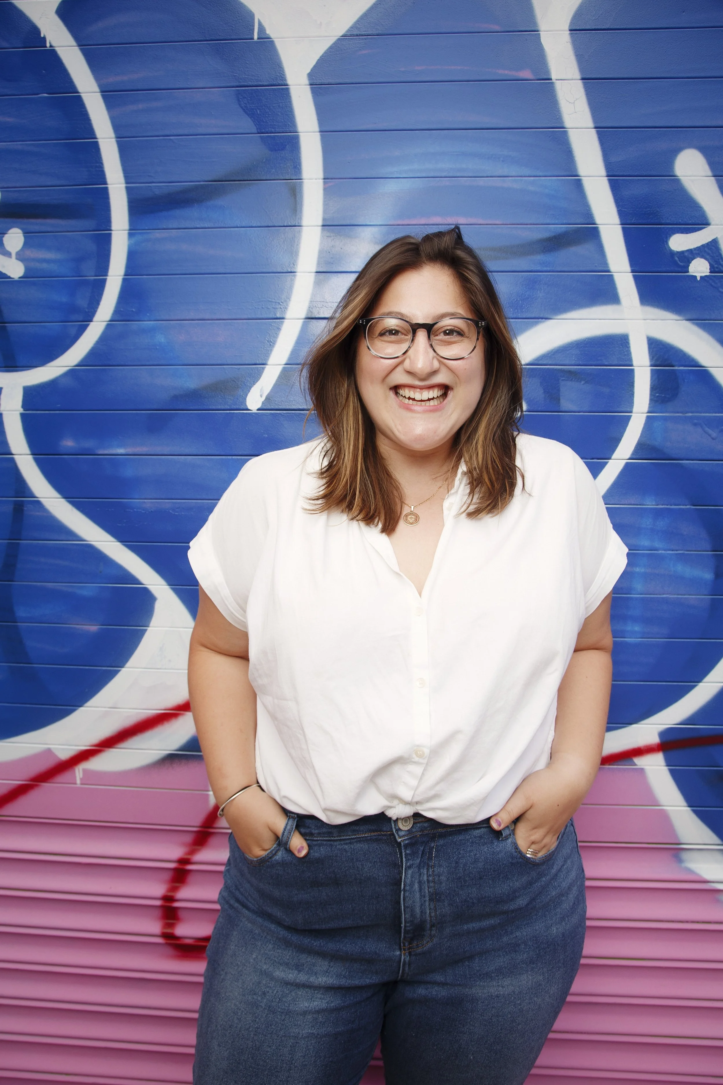 A young woman with brown hair, wearing glasses, a white button-up shirt, and jeans, smiling and standing with her hands in her pockets in front of a colorful graffiti wall with blue and pink hues.
