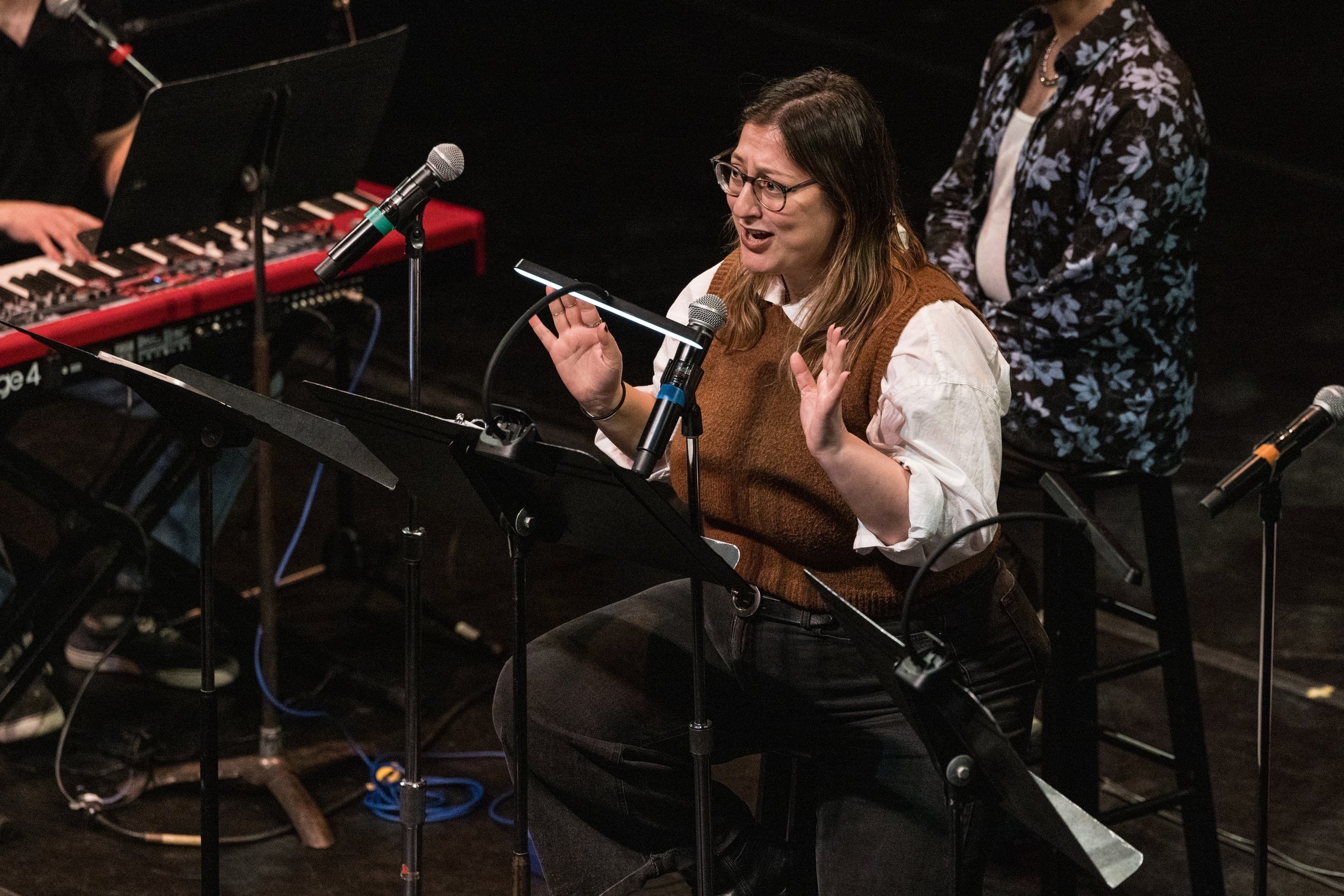 A woman with glasses and long brown hair singing into a microphone, with her hands raised, sitting at a music stand in a dimly lit setting. A woman in a floral shirt is visible in the background, and part of a keyboard and microphone setup are also v