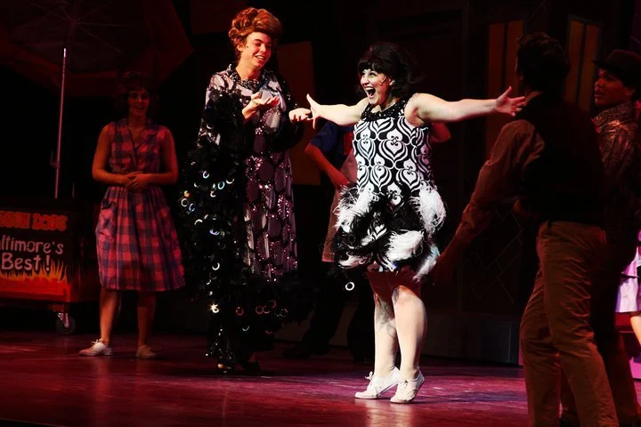 A theatrical stage scene with five women in colorful costumes. Two women in the center are smiling and showing expressive gestures, wearing patterned dresses. Other women are standing around them, with one in a pink checkered dress. The background ha
