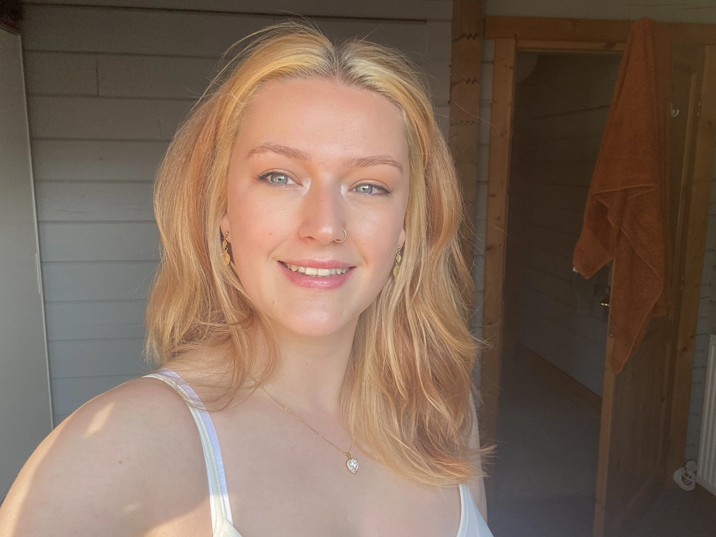 A young woman with blonde hair and blue eyes smiling outdoors, wearing a white tank top, jewelry, and a septum piercing.