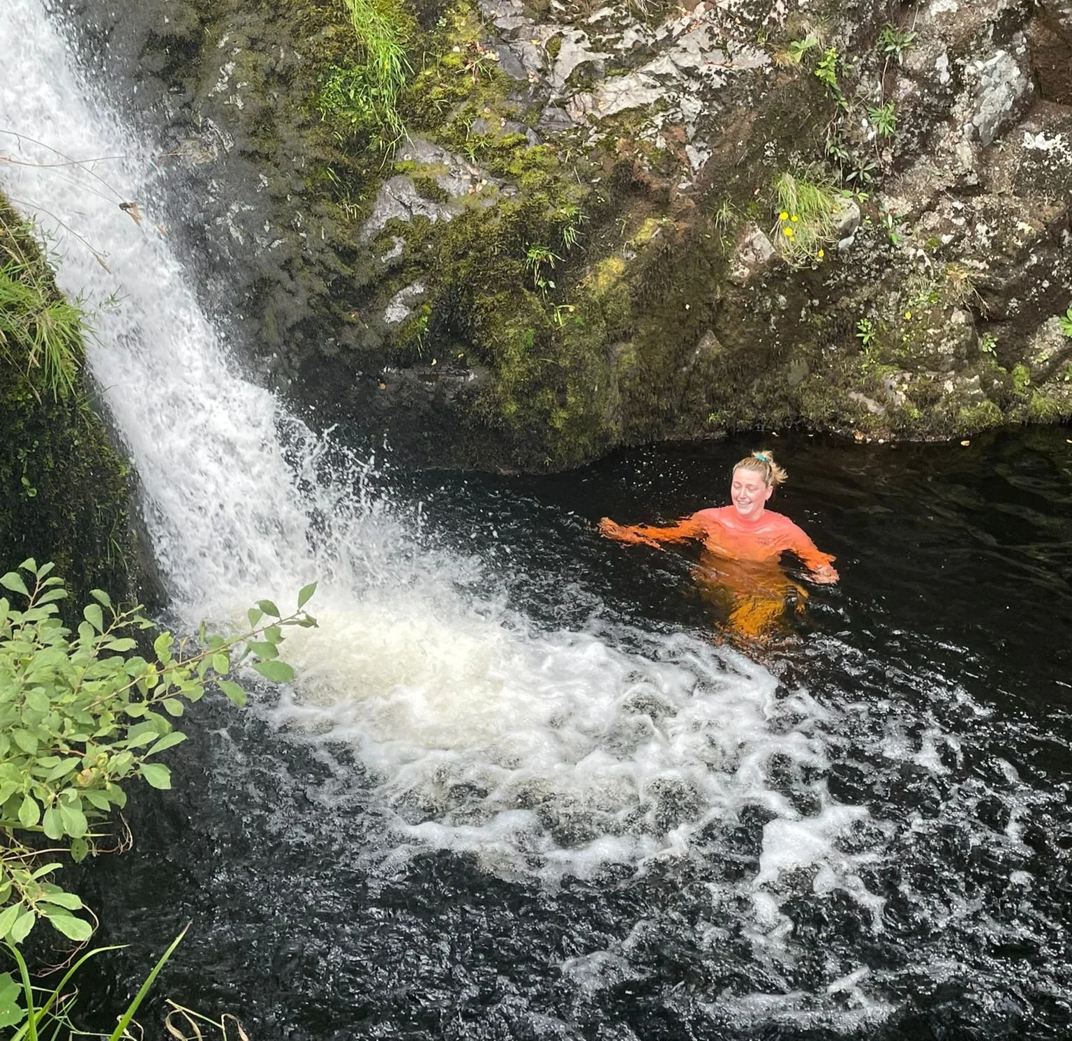 A woman swimming near a small waterfall in a natural, rocky setting with greenery.