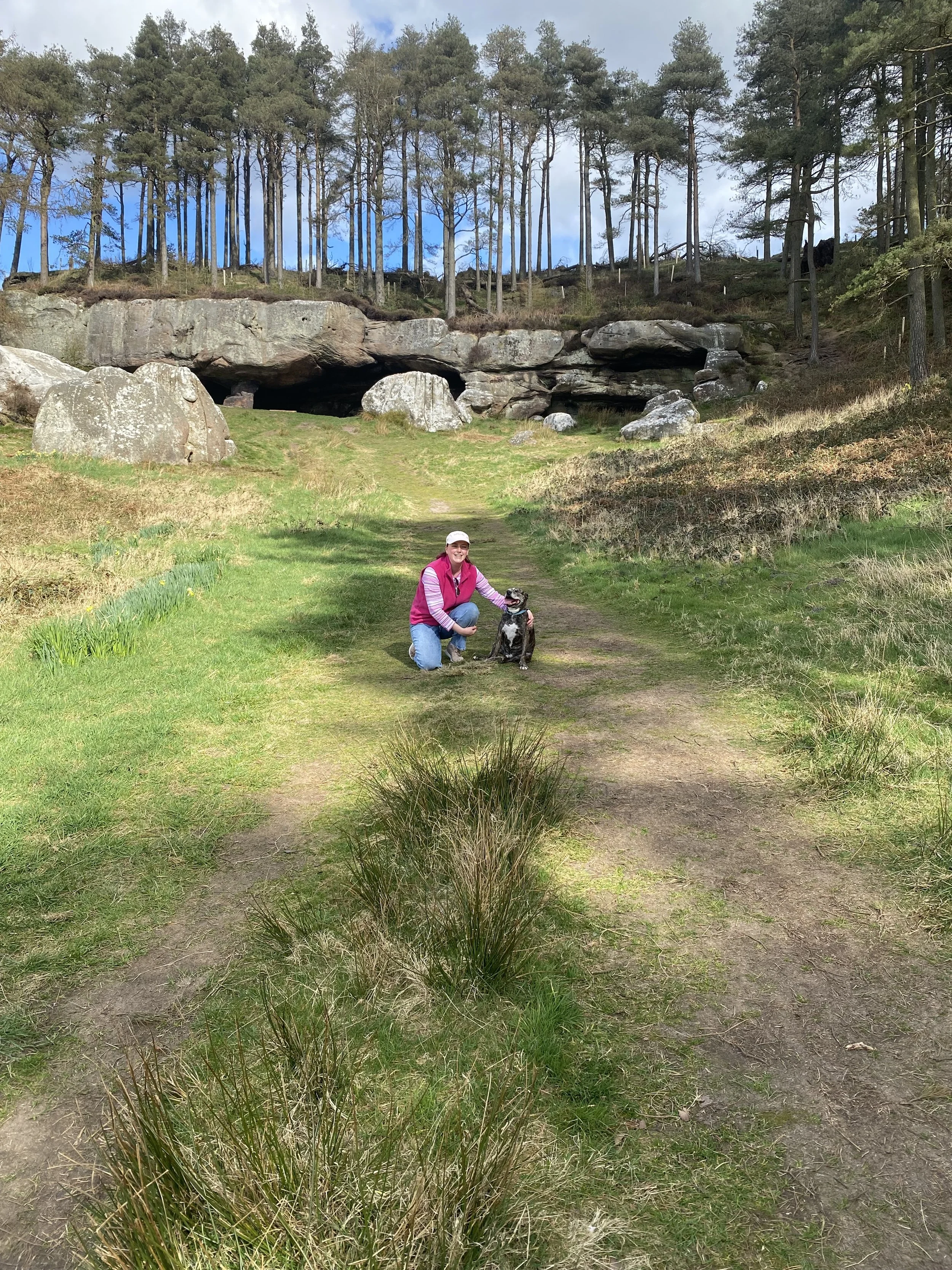A woman in a pink vest and gray hat kneeling on a trail with a black and white dog in front of a rocky hillside with a cave opening, surrounded by tall pine trees and lush green grass.