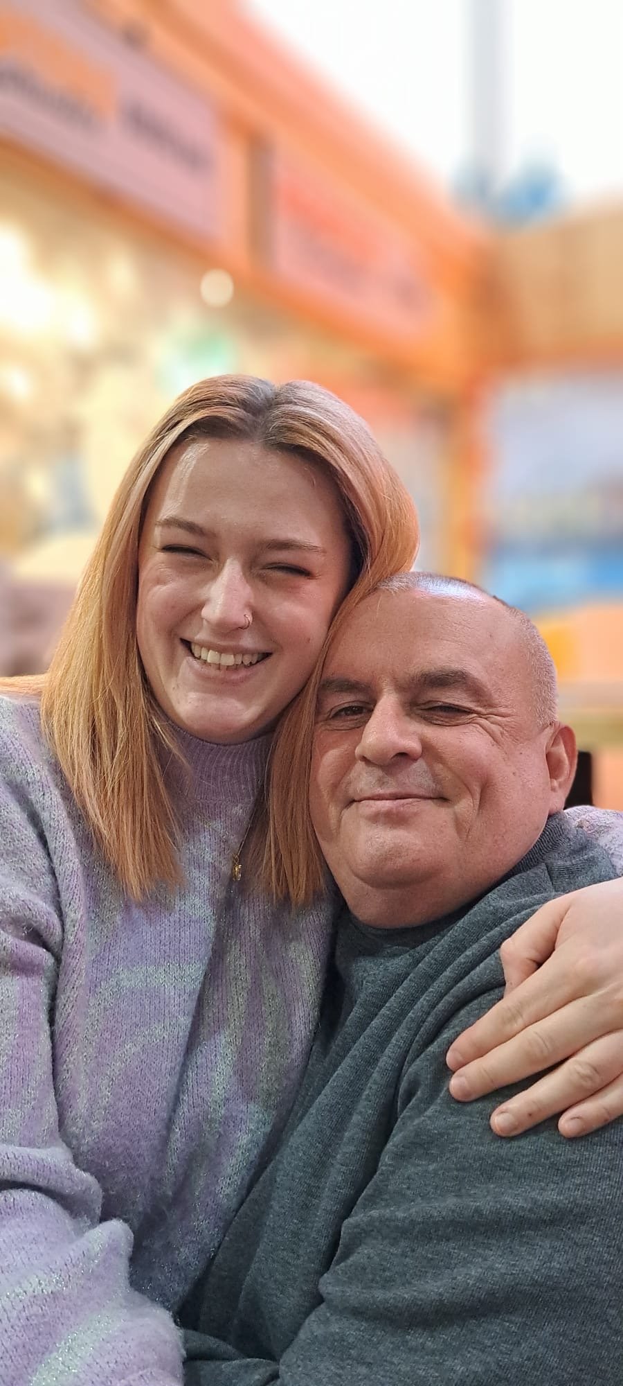A young woman with red hair and a nose ring smiling and hugging a bald man with a slight smile in a casual setting.