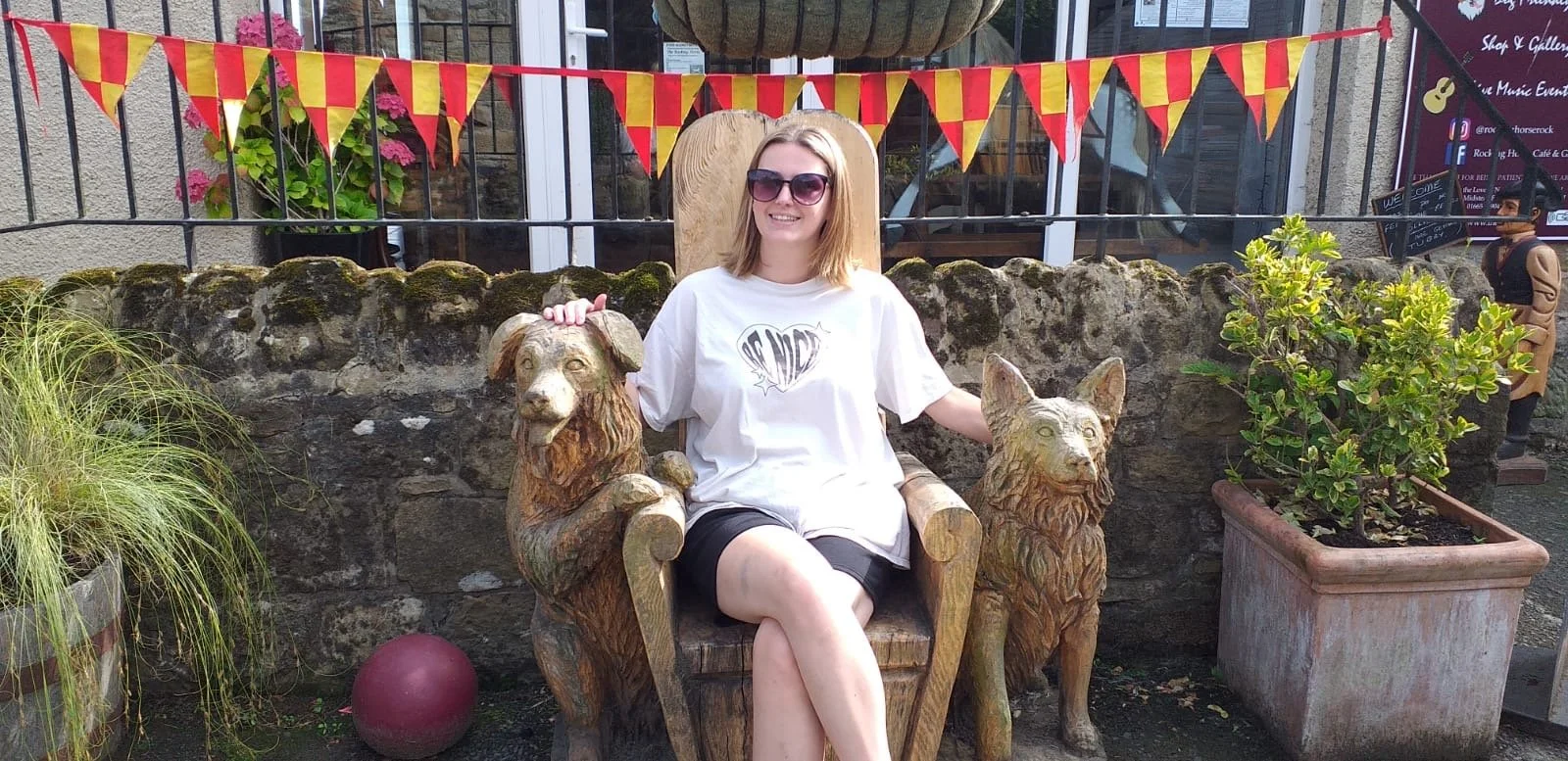 A woman wearing sunglasses and a white t-shirt sitting on a wooden throne-like chair with lion and wolf carvings, outdoors at a festive event with red and yellow bunting overhead and potted plants.