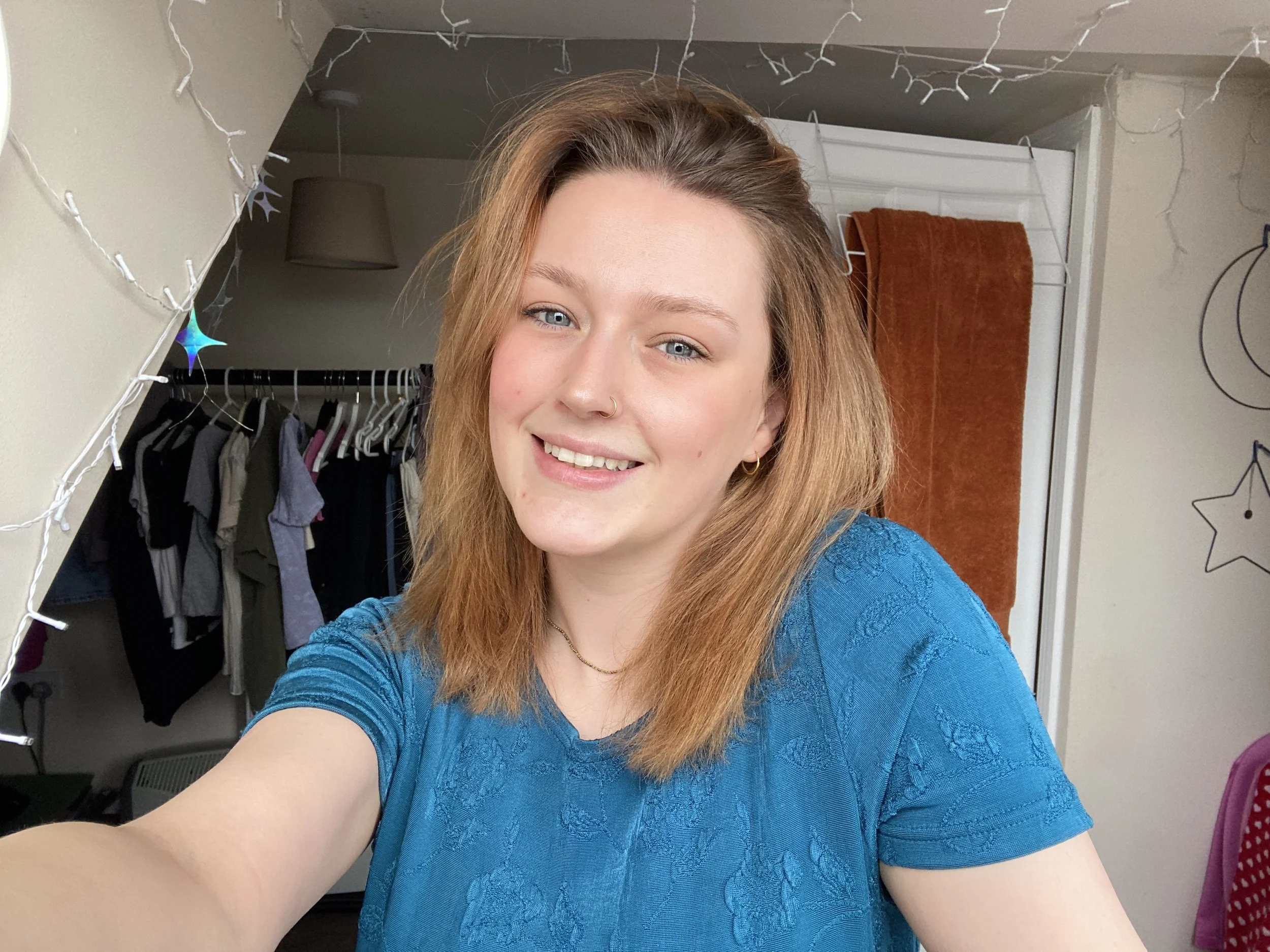 A young woman taking a selfie in her bedroom, smiling, with a closet and wall decorations visible in the background.