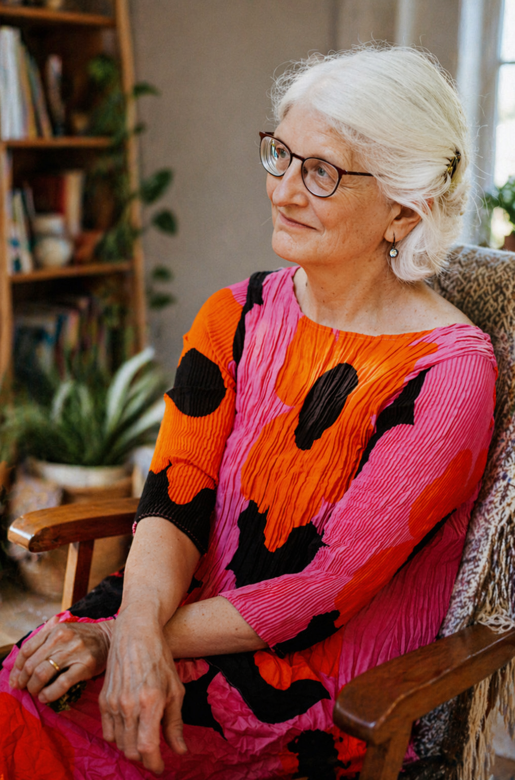 An elderly woman with white hair and glasses sitting in a rocking chair indoors. She wears a vibrant, colorful dress with abstract patterns in pink, orange, and black. There are bookshelves and plants in the background.