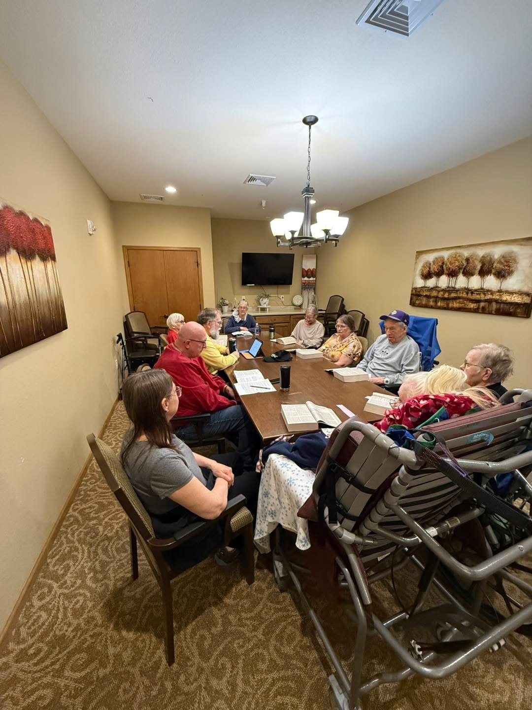 A group of elderly people seated around a rectangular dining table in a conference room, some reading, others listening, with a TV and artwork on the walls.