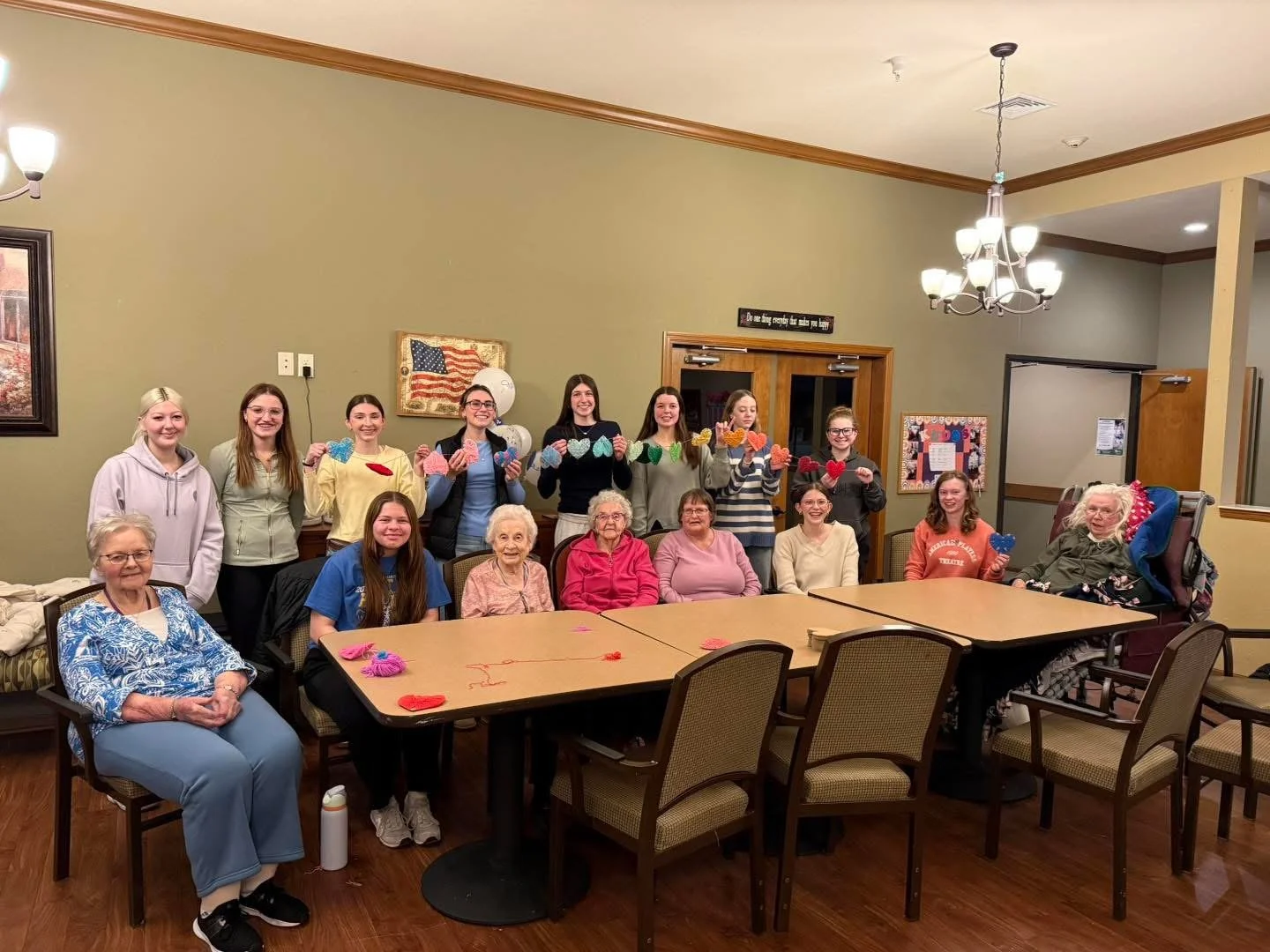 A group photo of women and girls, some elderly, in a room with green walls and wooden trim, holding colorful paper hearts. The group includes women sitting and standing, some holding handmade heart crafts, celebrating in a decorated room with balloons and framed artwork.