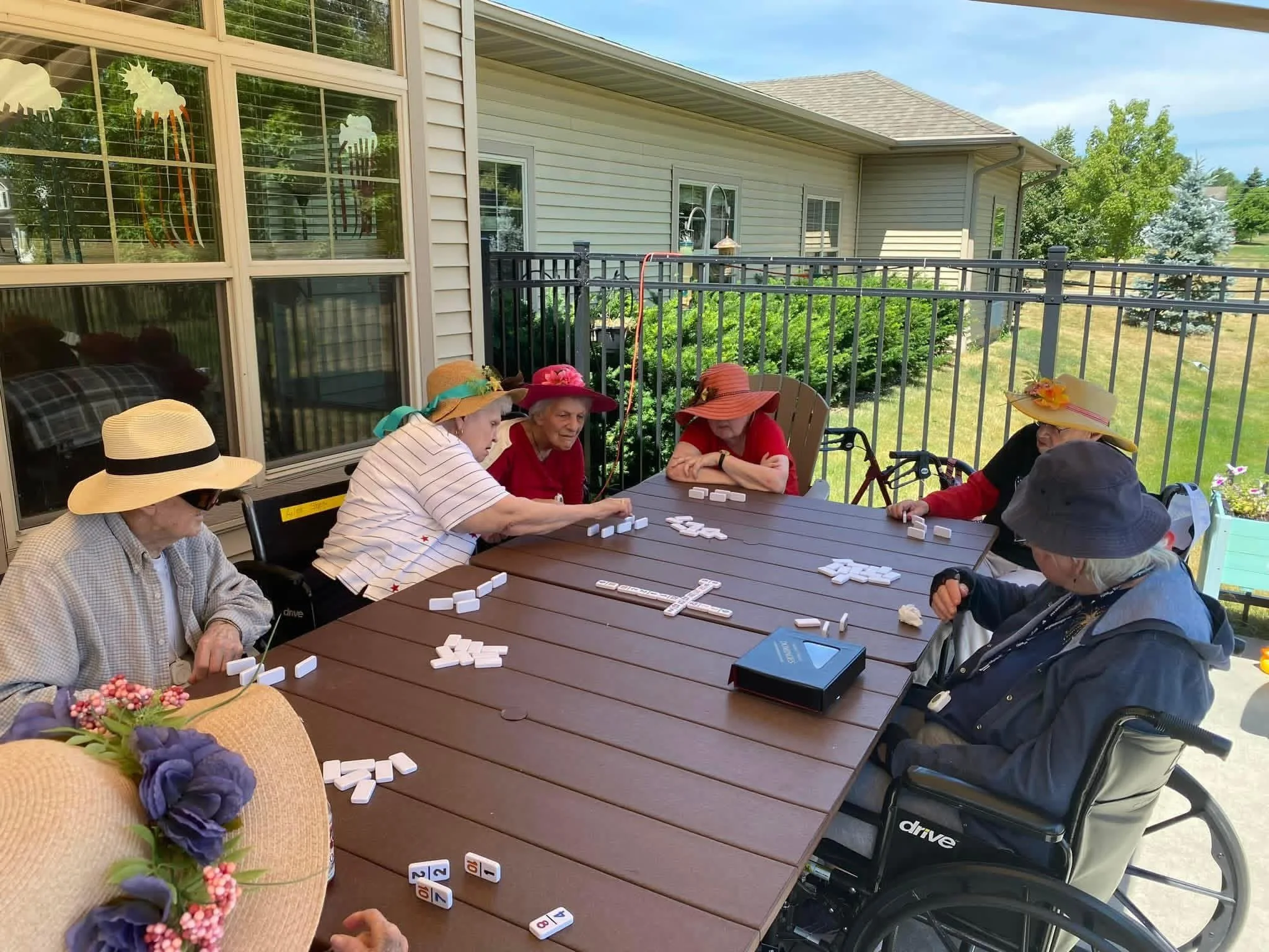 Group of elderly women playing lawn dominoes on a patio, wearing sun hats, with a house and trees in the background.