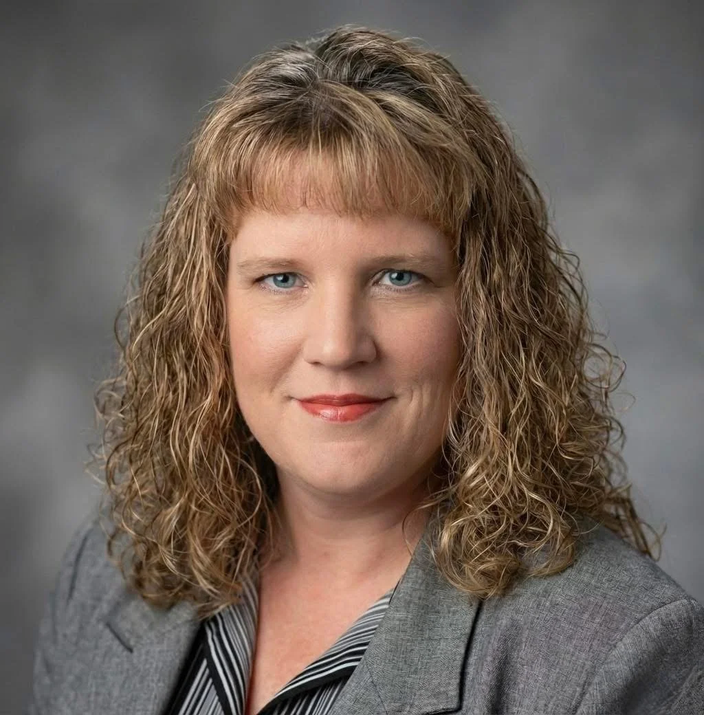 Professional headshot of a woman with curly blonde hair, wearing a grey blazer, smiling, against a grey background.