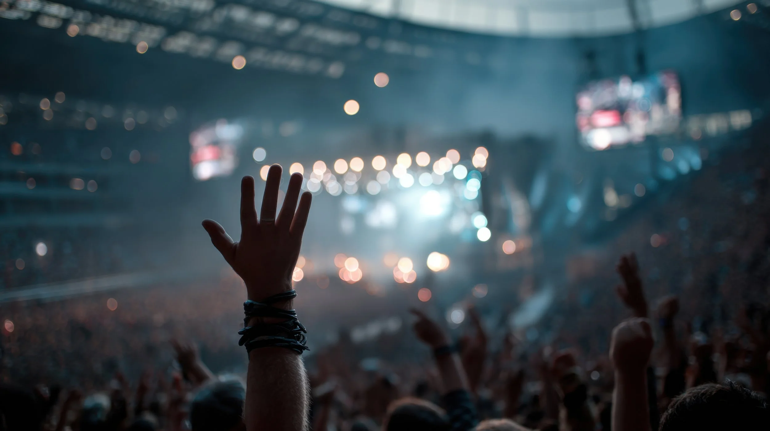 Person reaching out with their hand in a crowd at a concert or festival, with bright stage lights and blurred audience in the background.