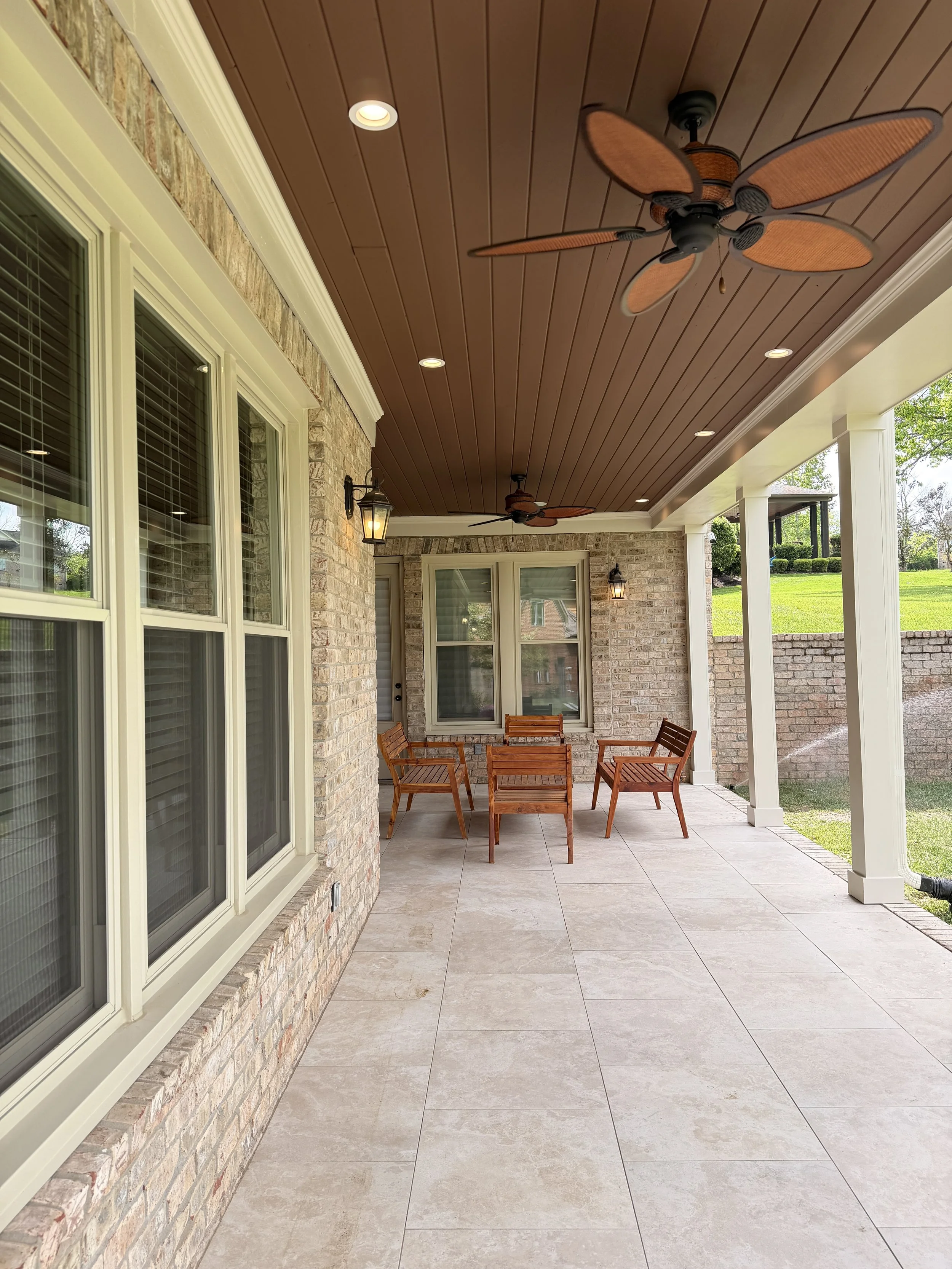 A covered outdoor patio area with a brick wall on one side, large windows, and a ceiling with wood paneling. The ceiling has two ceiling fans with leaf-shaped blades and recessed lighting. There are also wall-mounted lantern-style light fixtures. The