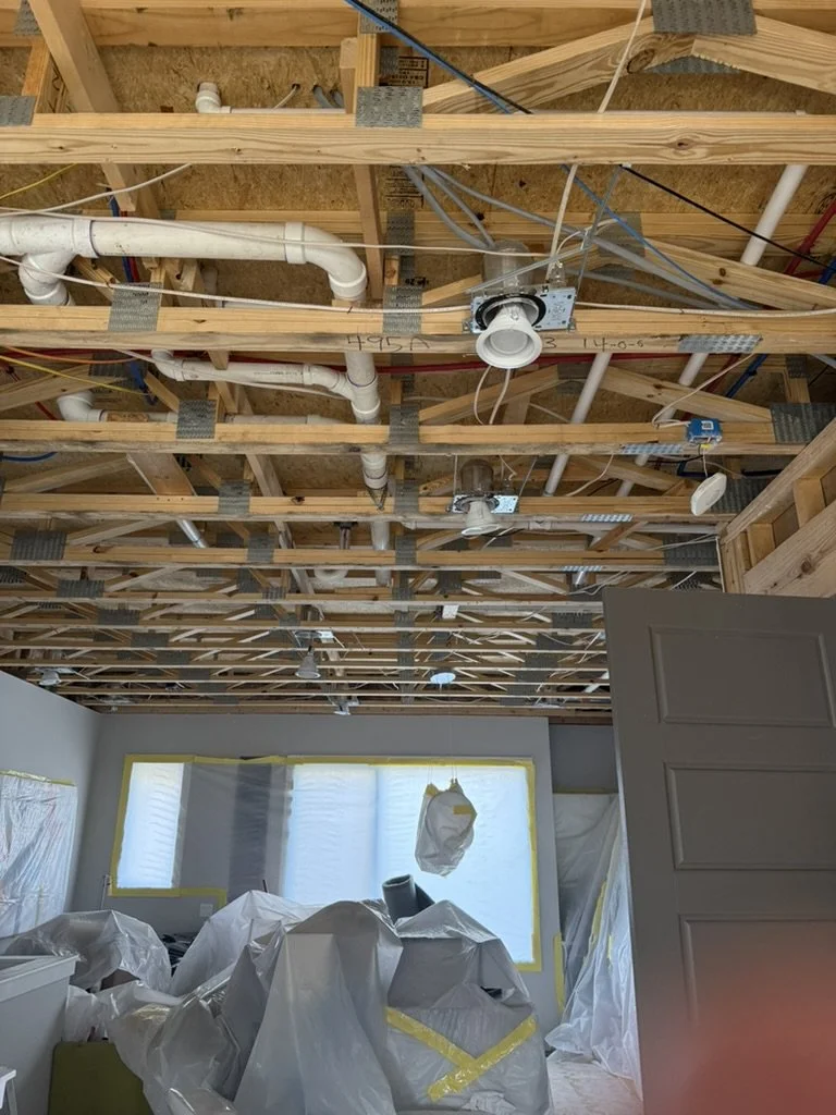 Interior of a room under construction with exposed wooden ceiling framework, visible pipes, wires, and electrical fixtures, and partially covered window and furniture.