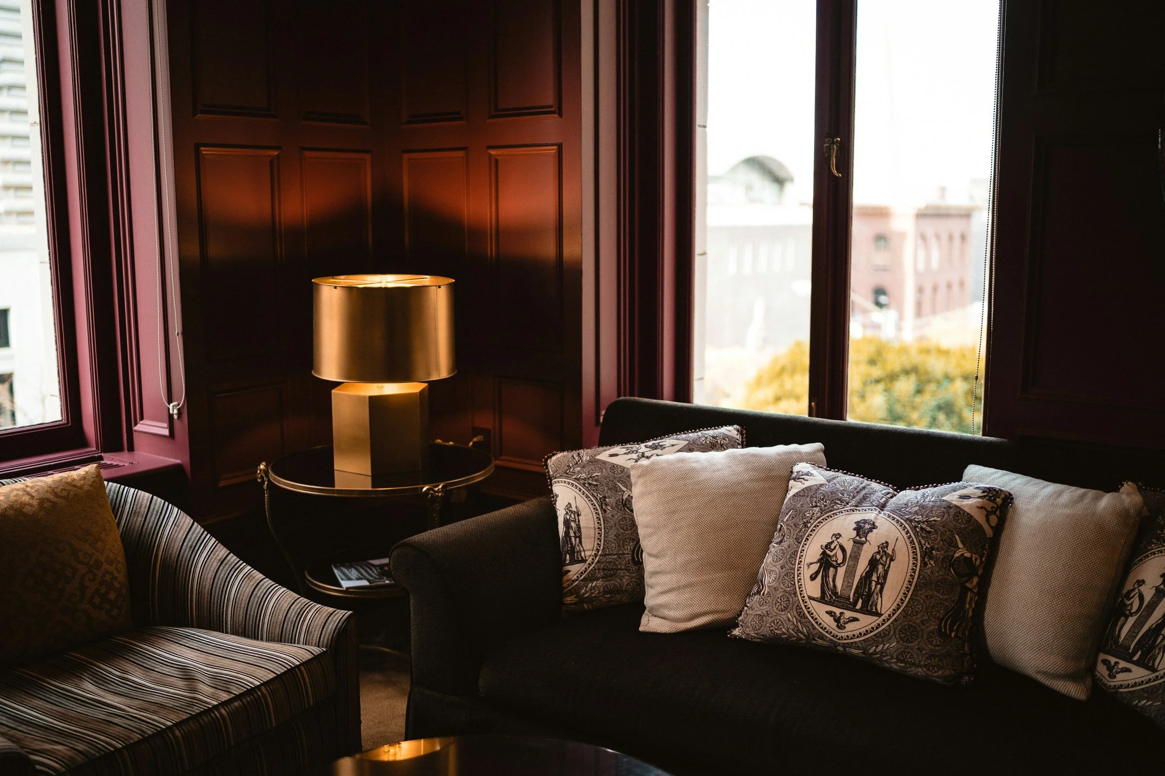 Interior of a room with dark wood paneling, large windows, a gold lampshade on a side table, and various patterned pillows on the sofa.