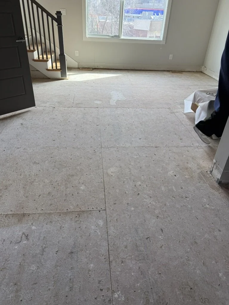 Empty room with unfinished flooring, a window letting in natural light, staircase with black railing at the left, and some construction materials covered on the right.