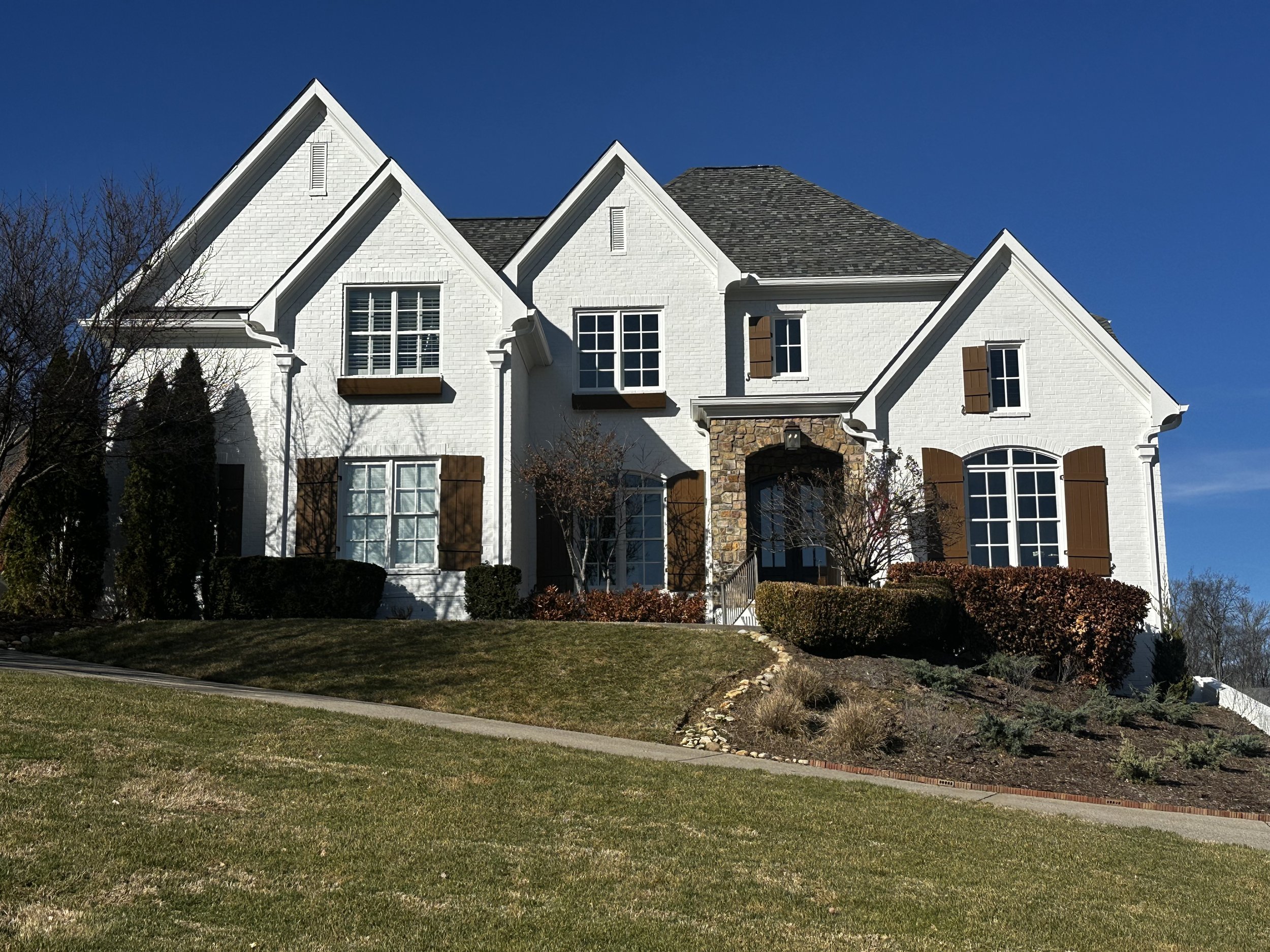 A white multi-story house with a gray shingle roof, multiple windows with shutters, and a stone archway entrance. The house is surrounded by a lawn and shrubbery, with some leafless trees in the yard, under a clear blue sky.