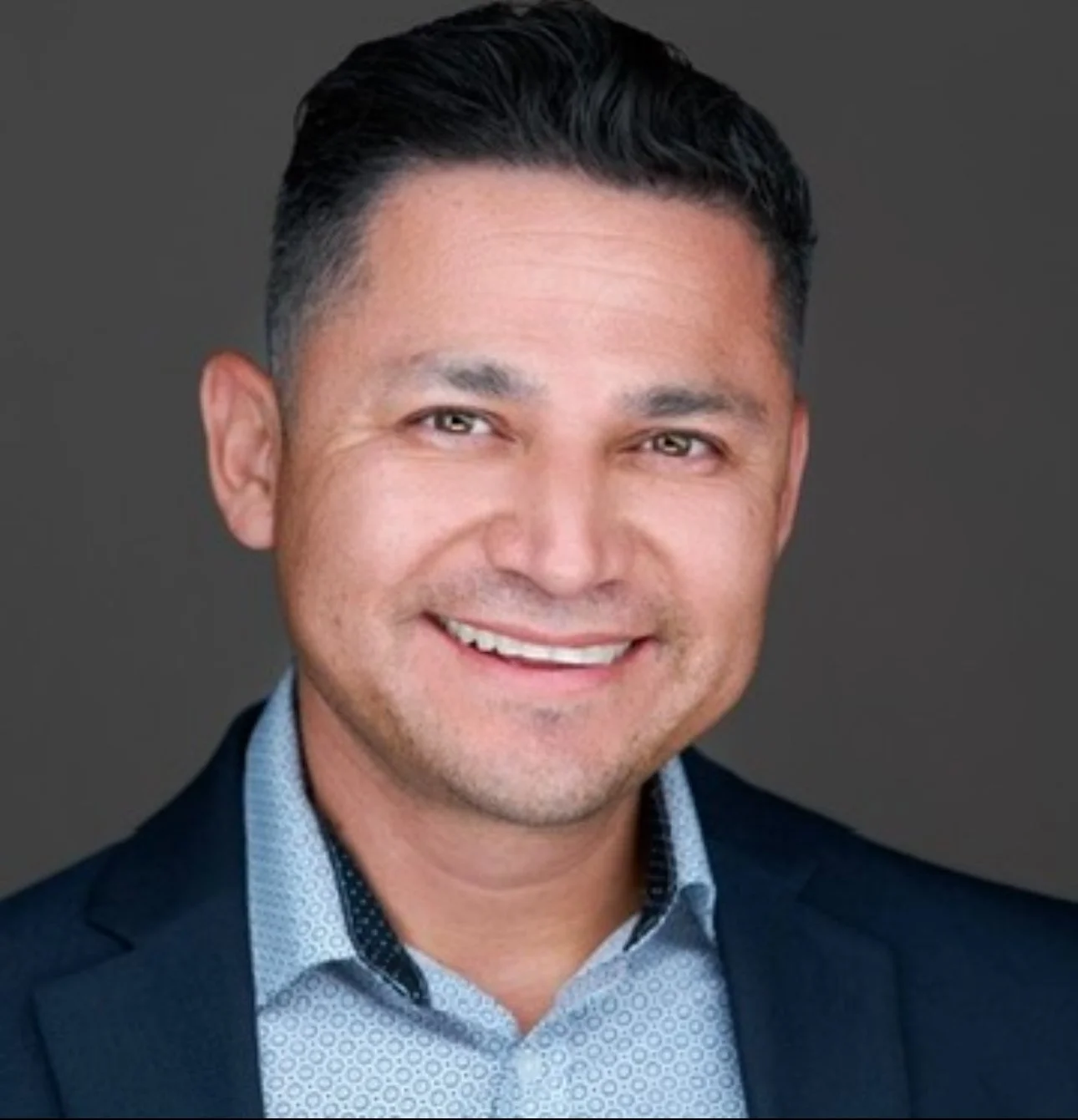 Headshot of a smiling man with short dark hair, wearing a navy blazer and a light patterned dress shirt, against a gray background.
