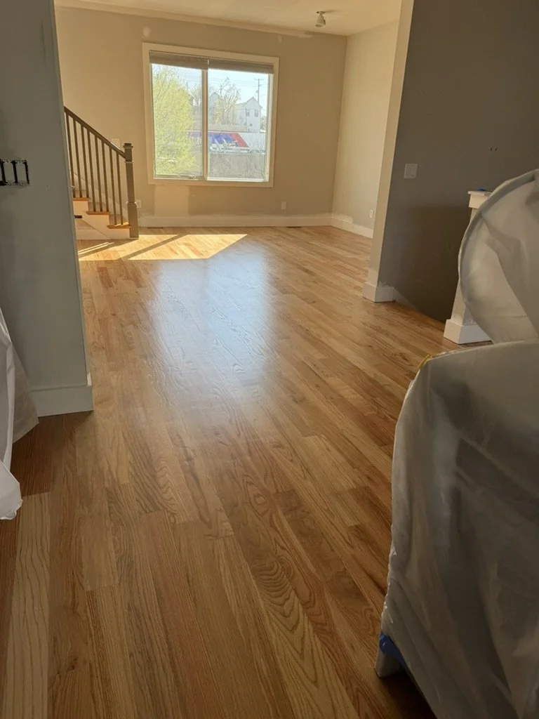 Empty living room with large window, hardwood flooring, staircase on the left, and a covered object on the right.