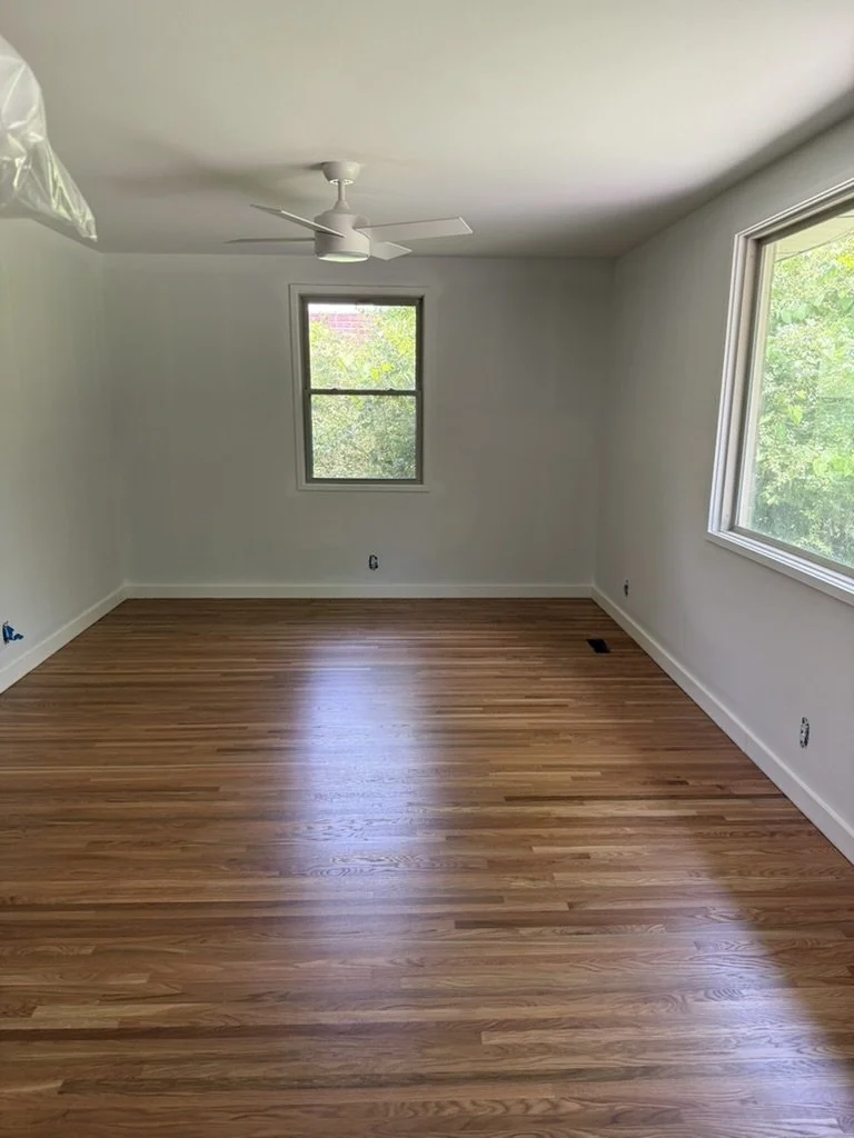 Empty room with white walls, hardwood floor, a ceiling fan, and large windows showing green trees outside.