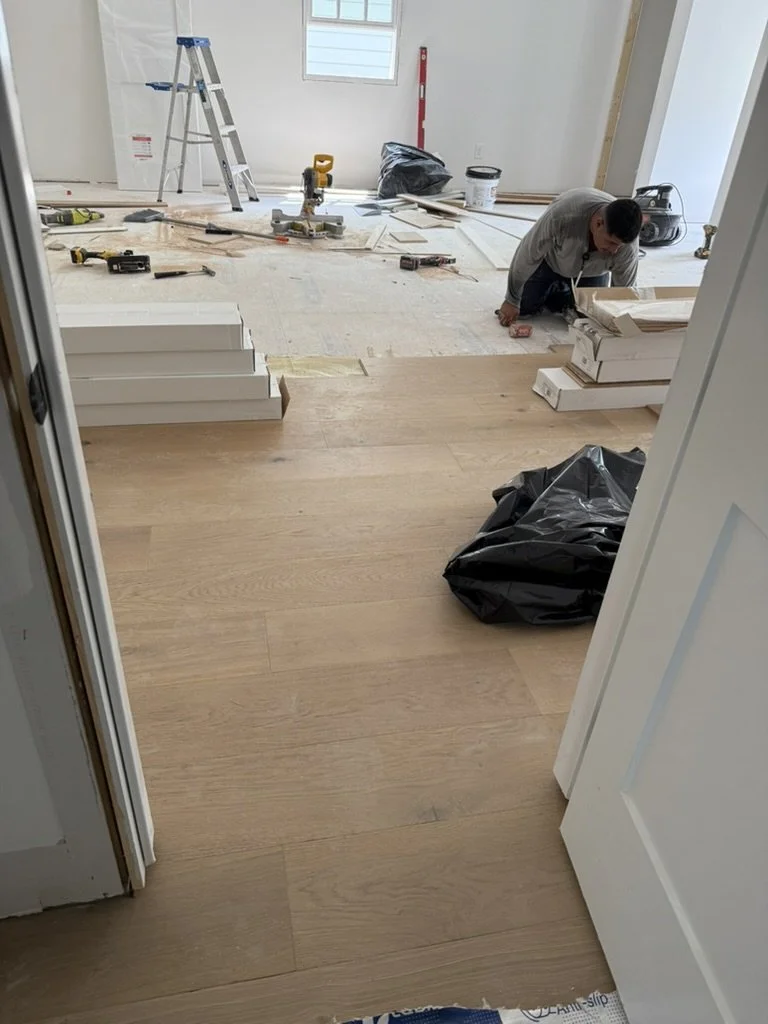 A person installing or working on hardwood flooring in a room under construction with tools, ladder, and construction materials scattered around.