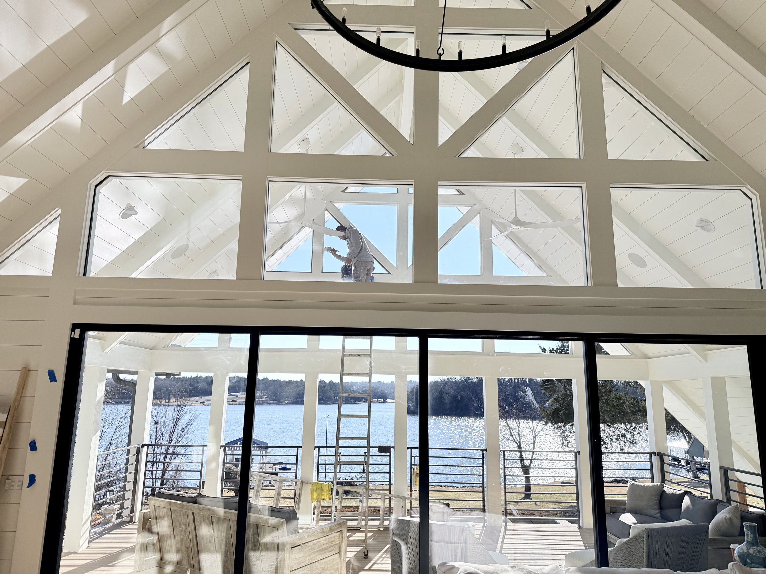 A person on a ladder cleaning large windows inside a bright room with a chandelier and a view of a lake and trees outside.
