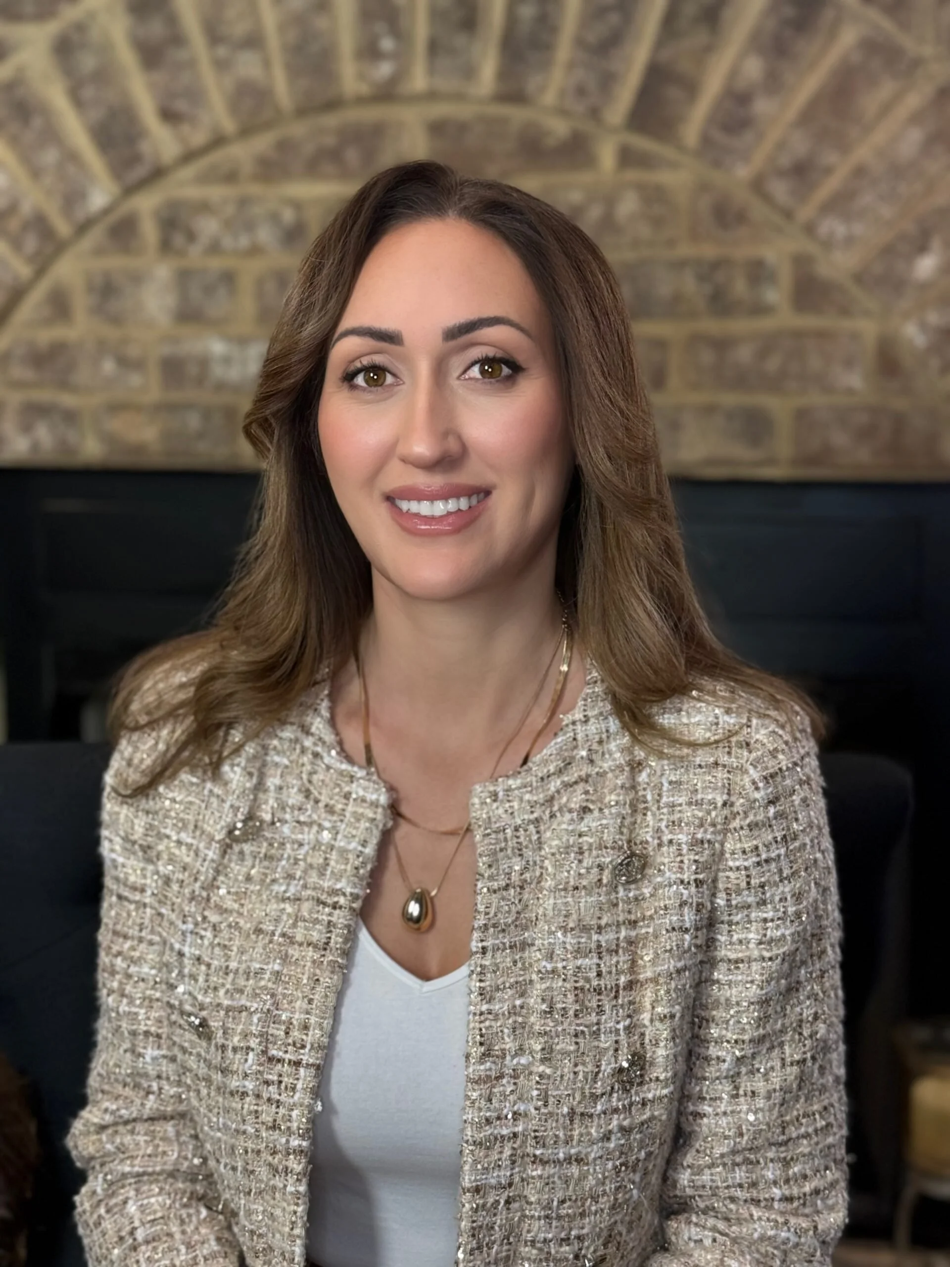 A woman with light brown hair smiling, wearing a textured beige blazer, a white top, and a gold and black necklace, sitting in front of a brick fireplace wall.