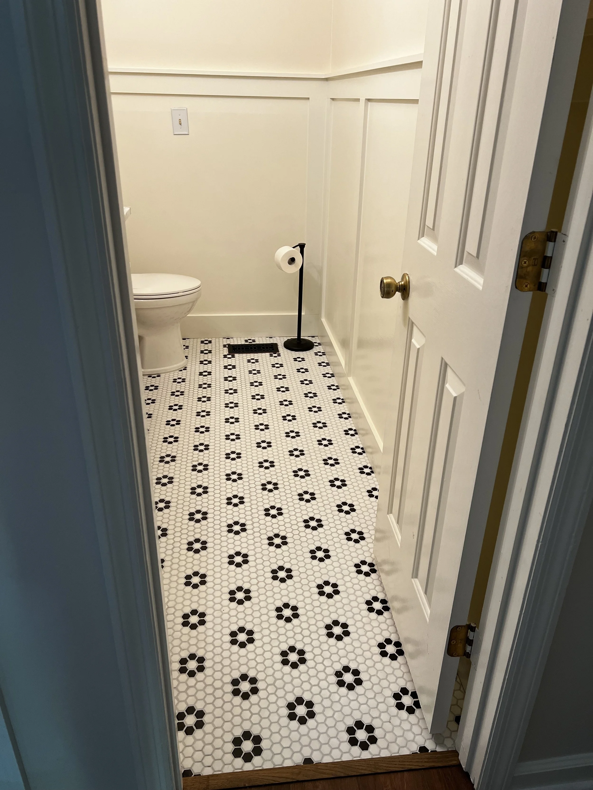 Inside a small bathroom, showing a white toilet, black and white patterned tile floor, a toilet paper stand with a roll of toilet paper, and a sealed door.
