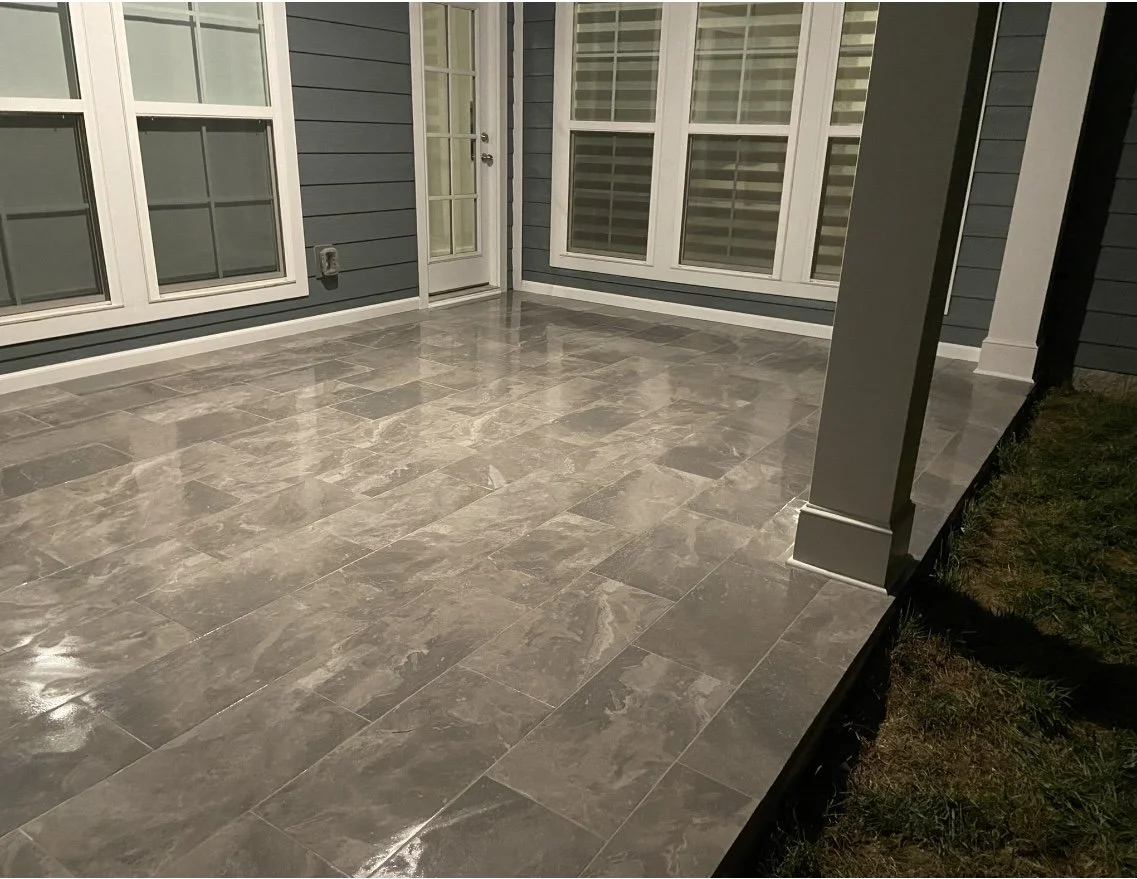 Empty outdoor patio with shiny gray tile flooring, blue house siding, white window frames, a glass door, and grass on the side.
