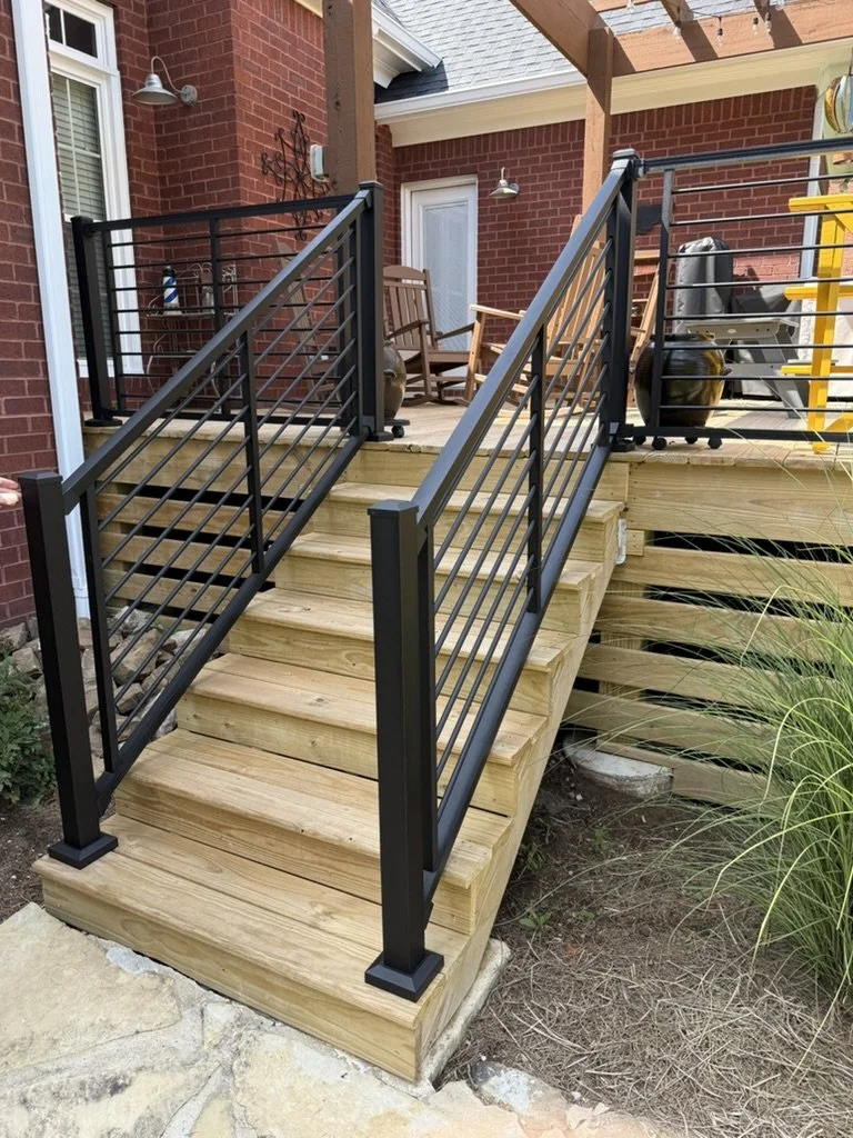 Newly built wooden deck with stairs and black metal railings attached to a brick house, featuring outdoor furniture and grilling equipment.