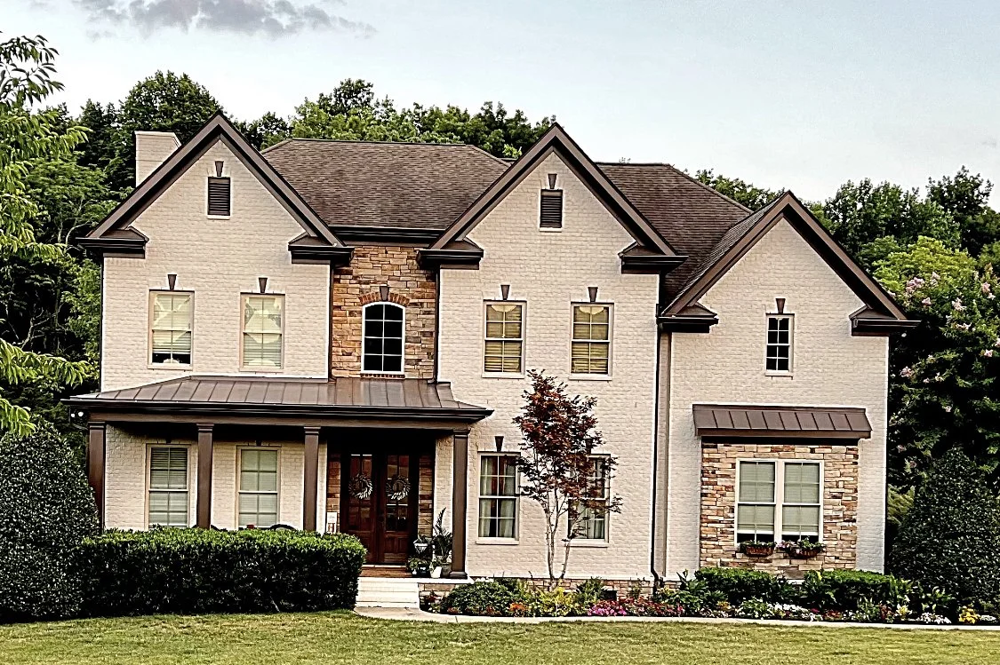 Large two-story house with a brick and siding exterior, brown roofing, and multiple windows, surrounded by green trees and lawn.