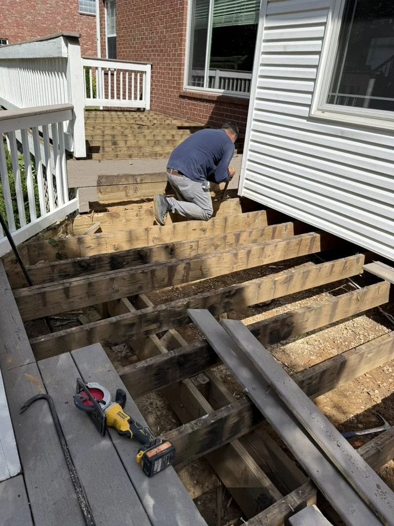 A person kneeling and working on building wooden stairs outside a house. The stairs are under construction, with some framing completed, and tools like a drill and hammer are visible on the porch.