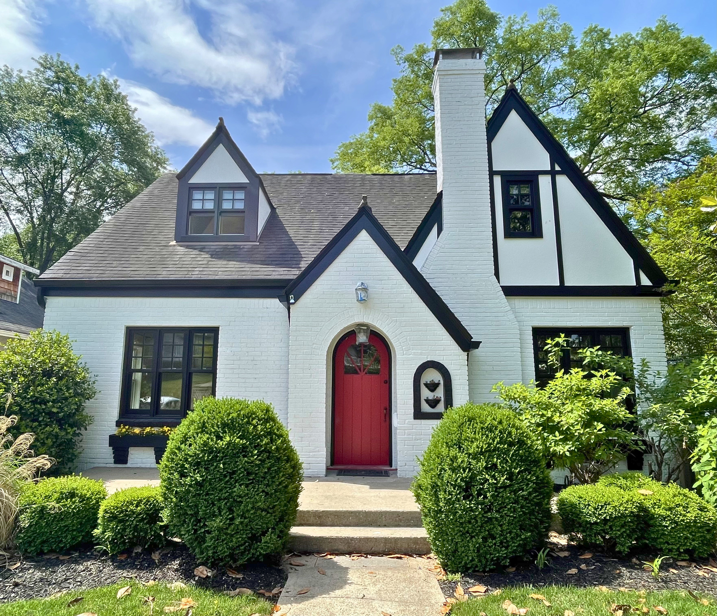 Front view of a white house with black trim and a red door, surrounded by green bushes and trees, under a blue sky with some clouds.