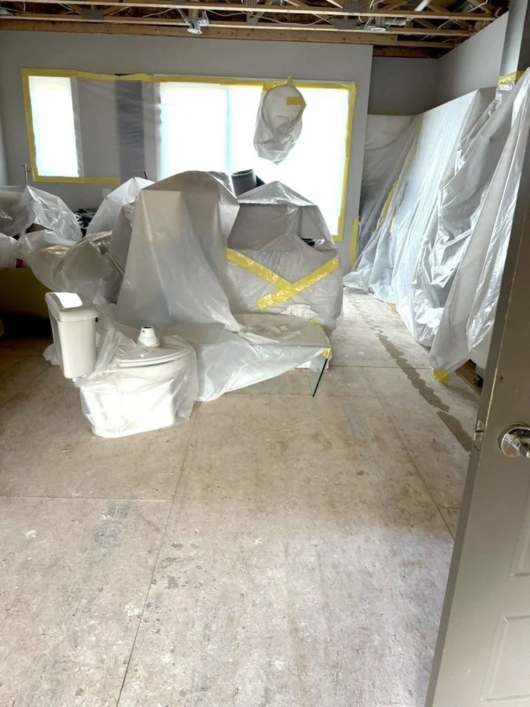 Room under construction with plastic sheeting covering furniture and walls, construction tape marking areas, and unfinished ceiling with exposed beams.