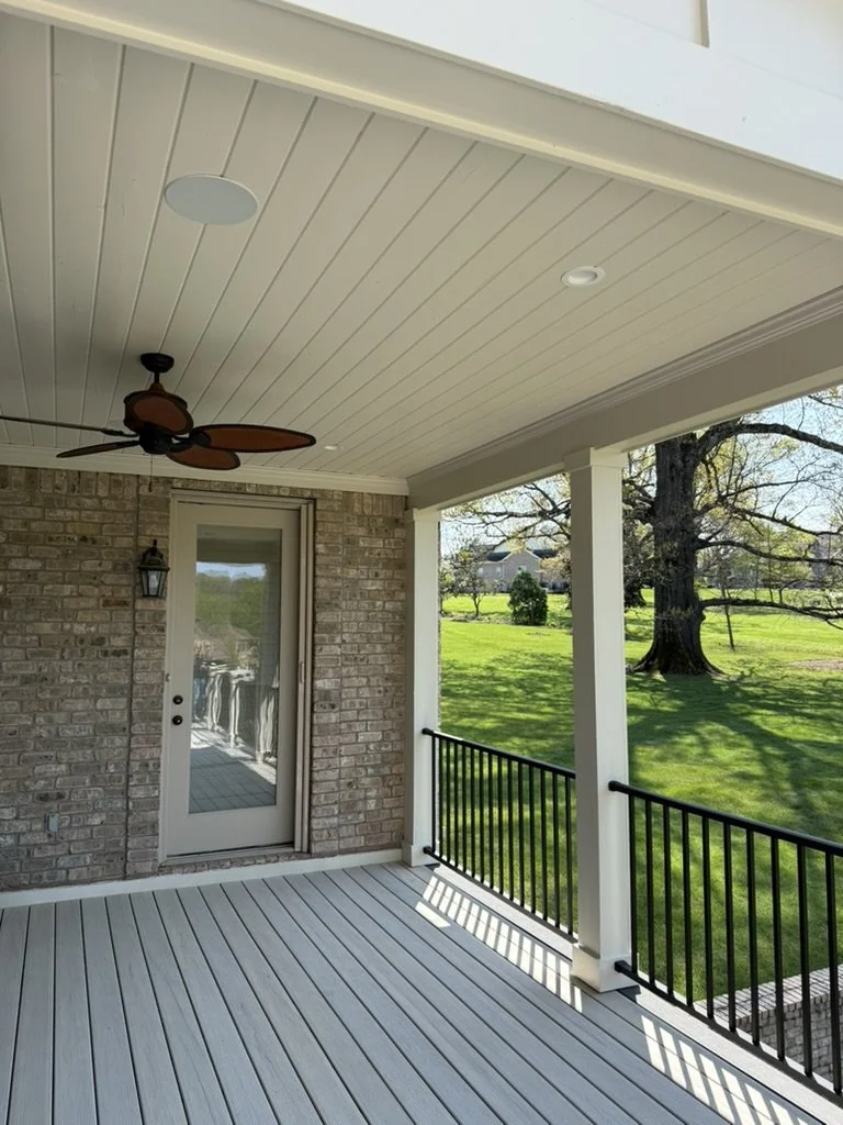 Covered porch with white decking, black railing, a ceiling fan, and a brick wall with a glass door leading outside, overlooking a green lawn and trees.