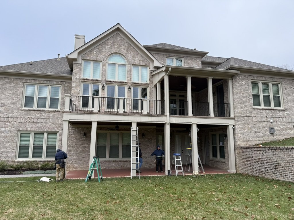 Workers installing a new balcony railing on the second-floor porch of a large brick house with multiple windows.