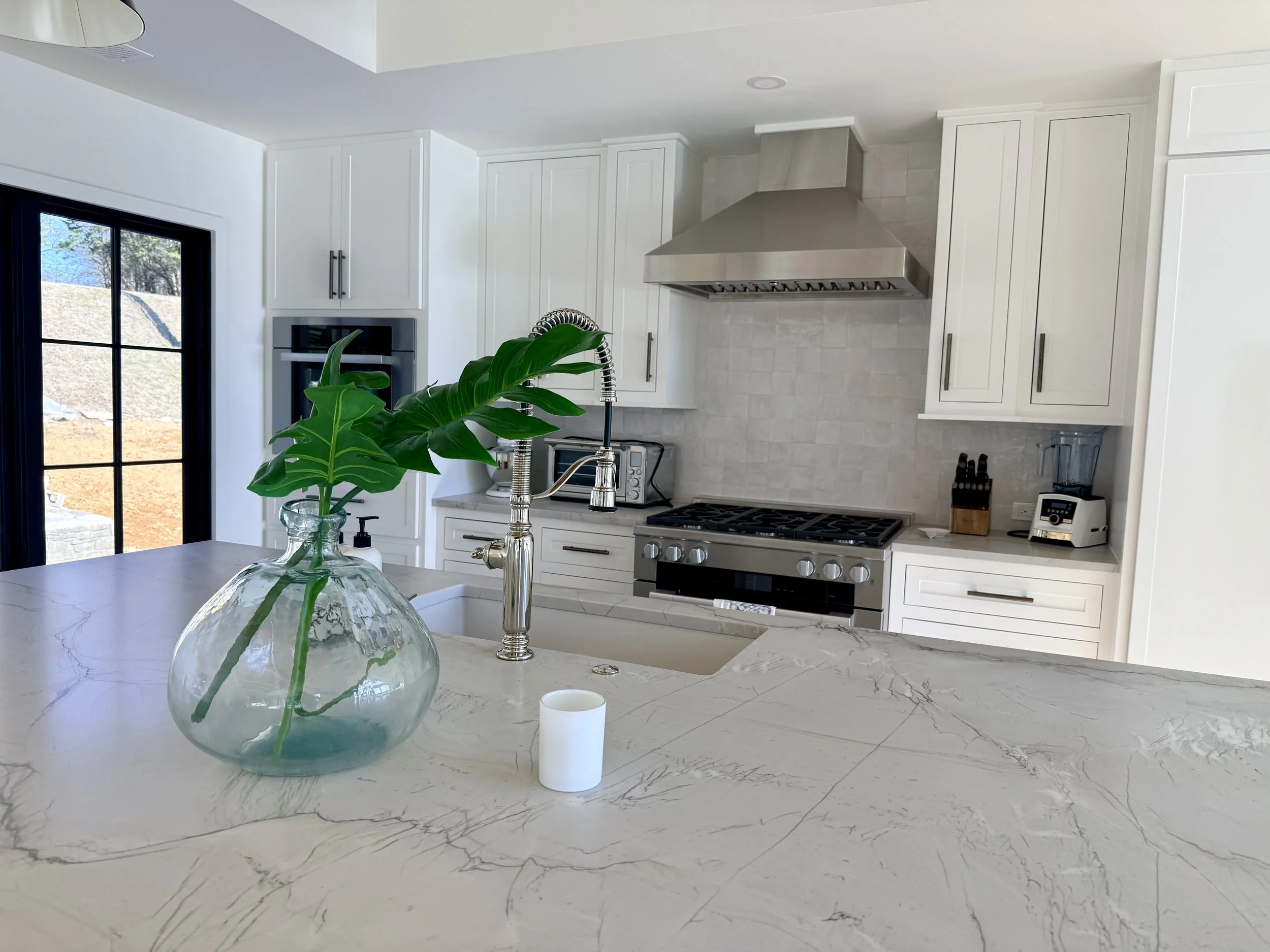 Modern kitchen with white cabinets, marble countertops, and a stainless steel range hood. A large window on the left shows an outdoor view. There is a green leafy plant in a glass vase on the counter, and small appliances including a blender and toas