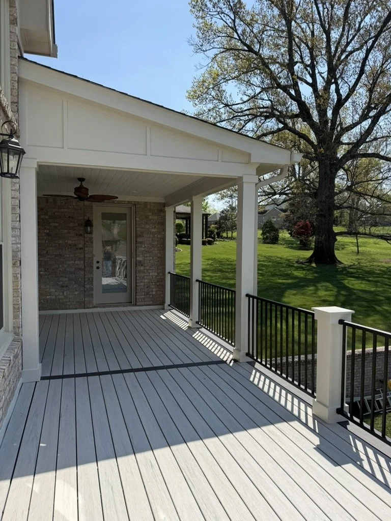 A spacious wooden porch with white railings and a black horizontal top rail. The porch is attached to a brick house with a glass door and a window. There is a ceiling fan and an outdoor lantern light. The porch overlooks a lush green yard with a larg