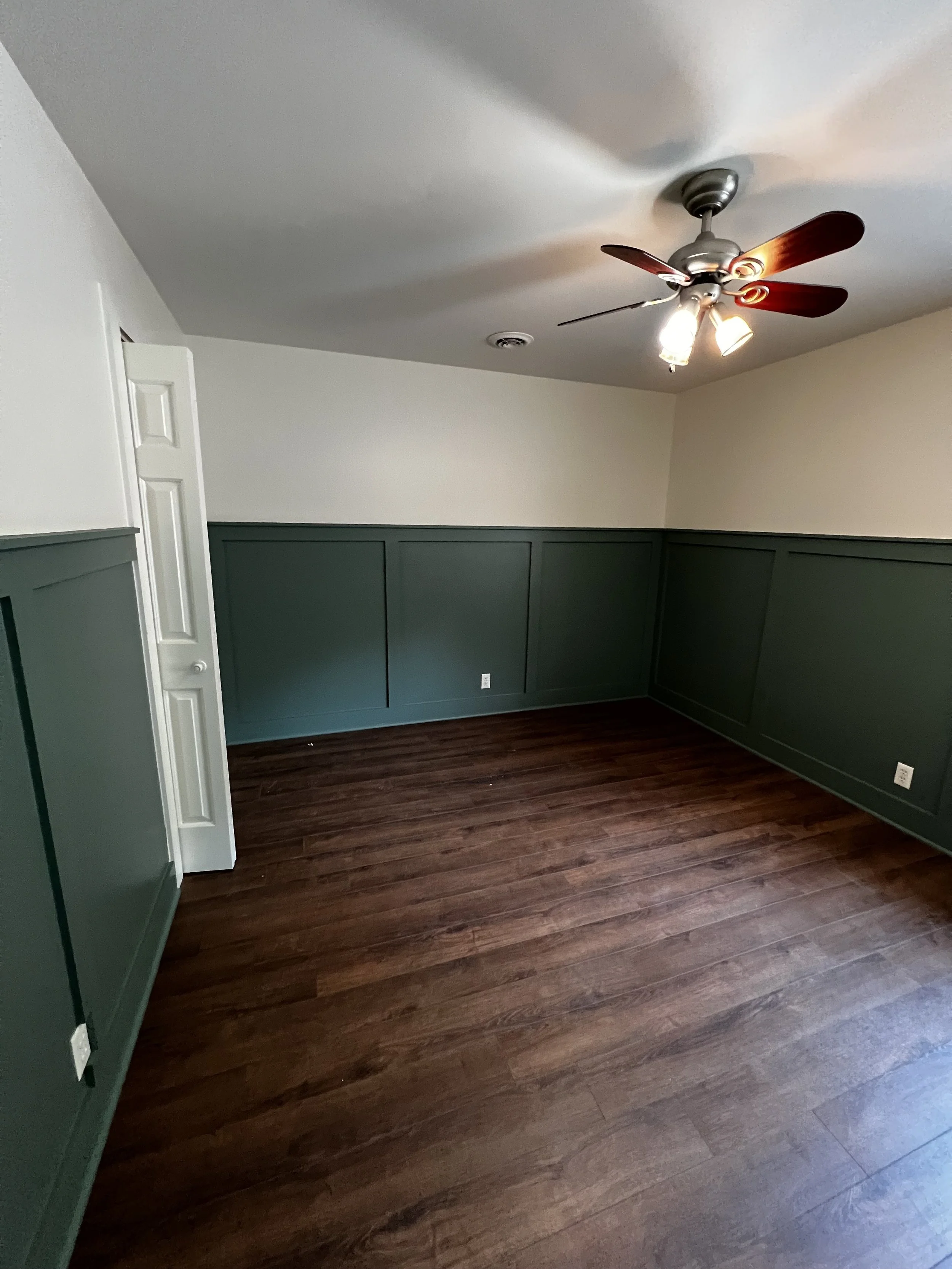Empty room with dark wood floors, green wainscoting, white walls, a ceiling fan with dark wooden blades and a light, and an open door.