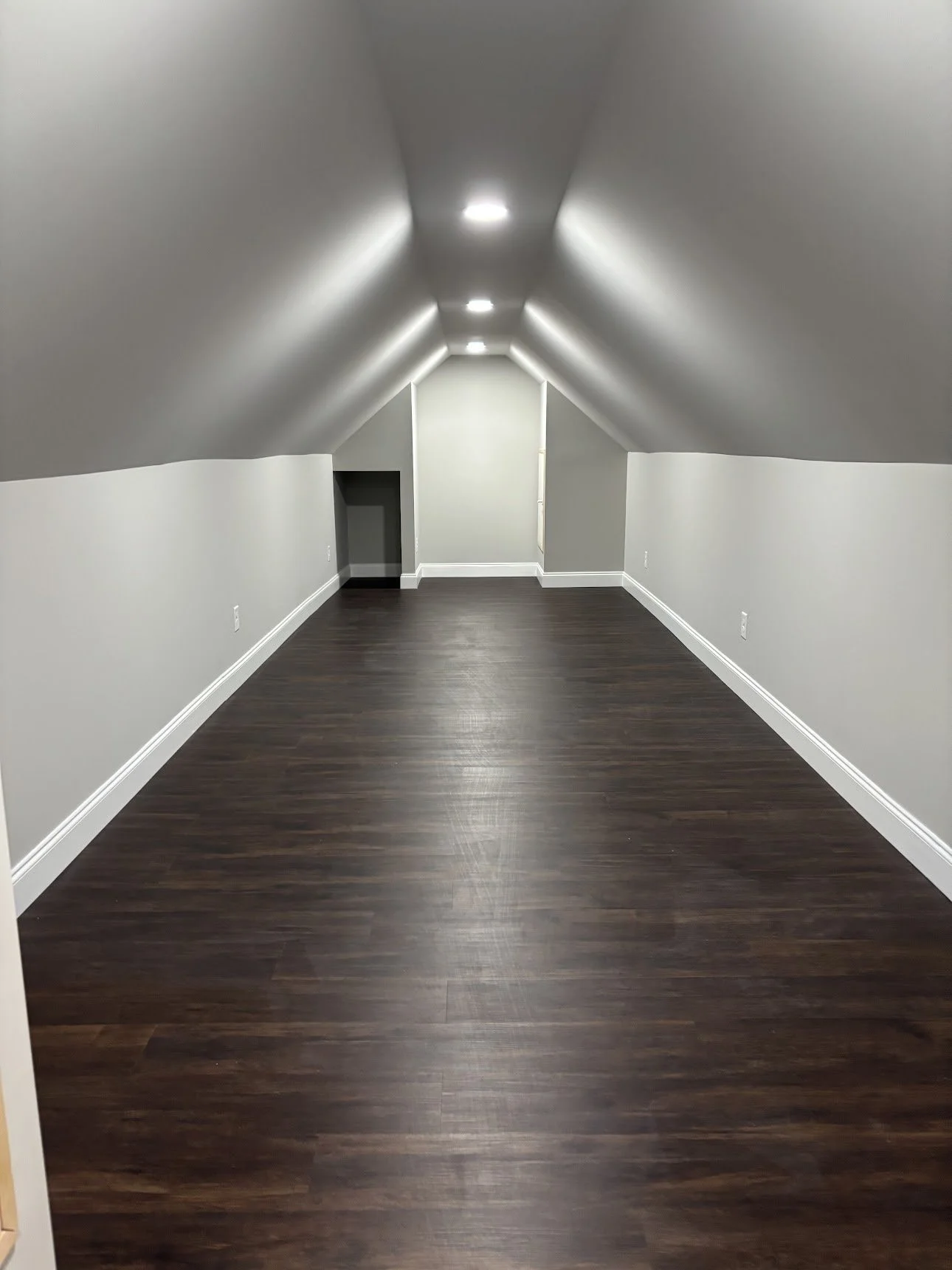 Empty finished attic with dark hardwood floor, white walls, sloped ceiling with recessed lights, and a small doorway on the left.