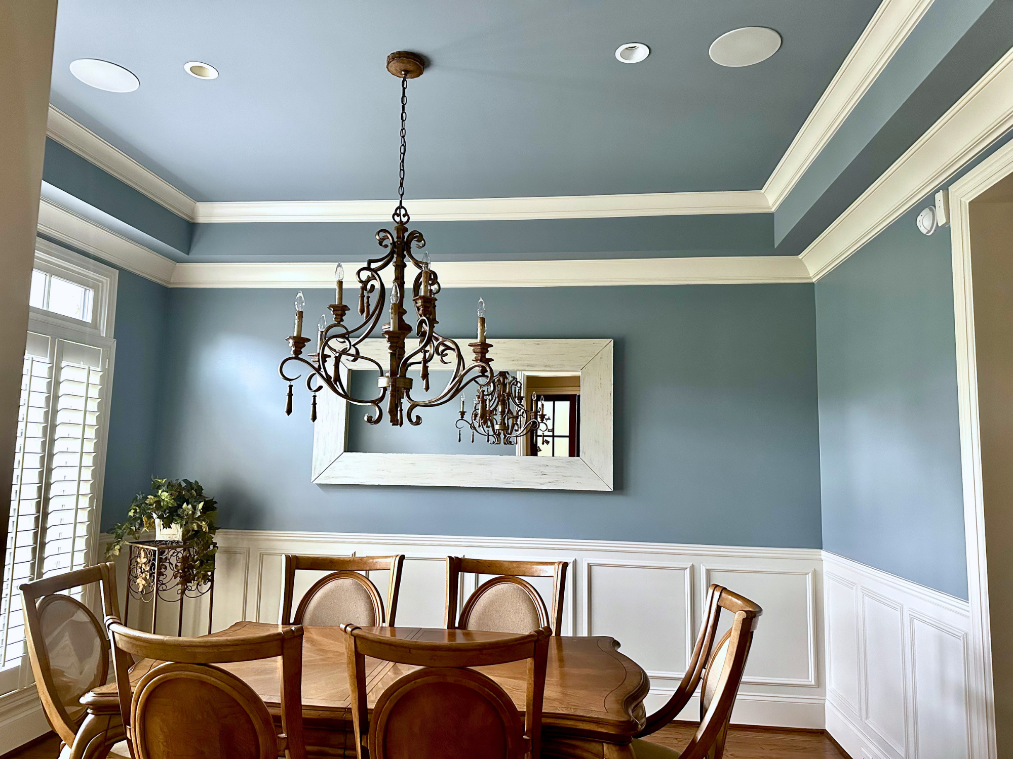 A dining room with a round wooden table, wooden chairs, decorative molding on the walls, a chandelier hanging from the ceiling, a large mirror, and a window with plantation shutters.