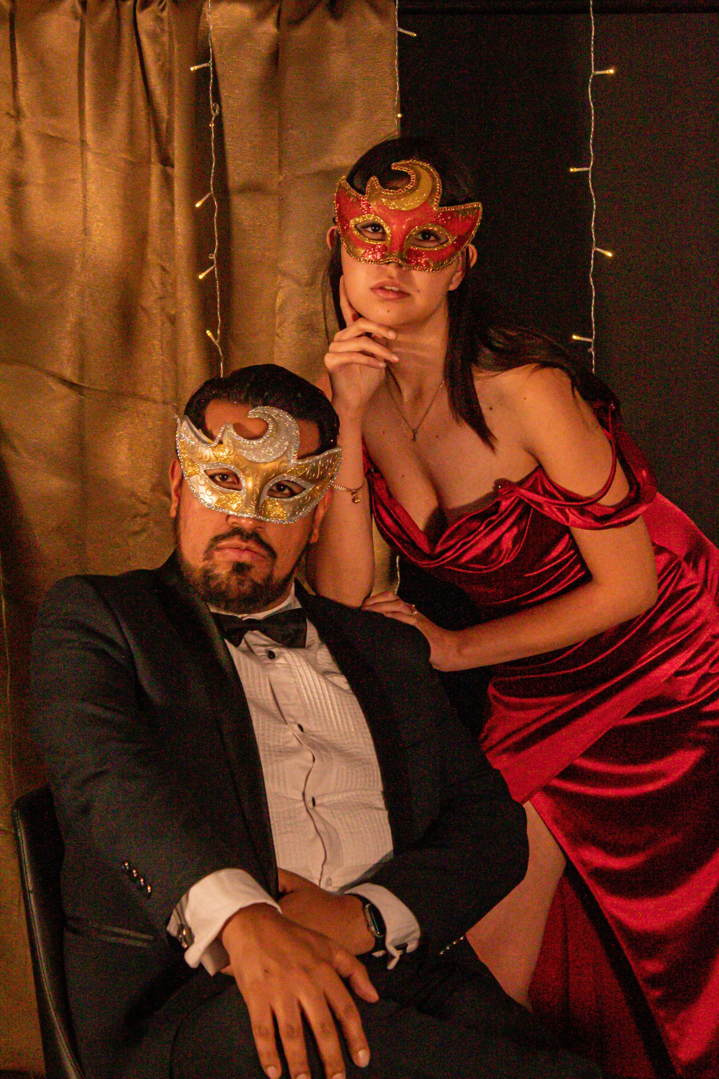 A man in a tuxedo and a woman in a red satin dress and masks celebrating at a masquerade party.