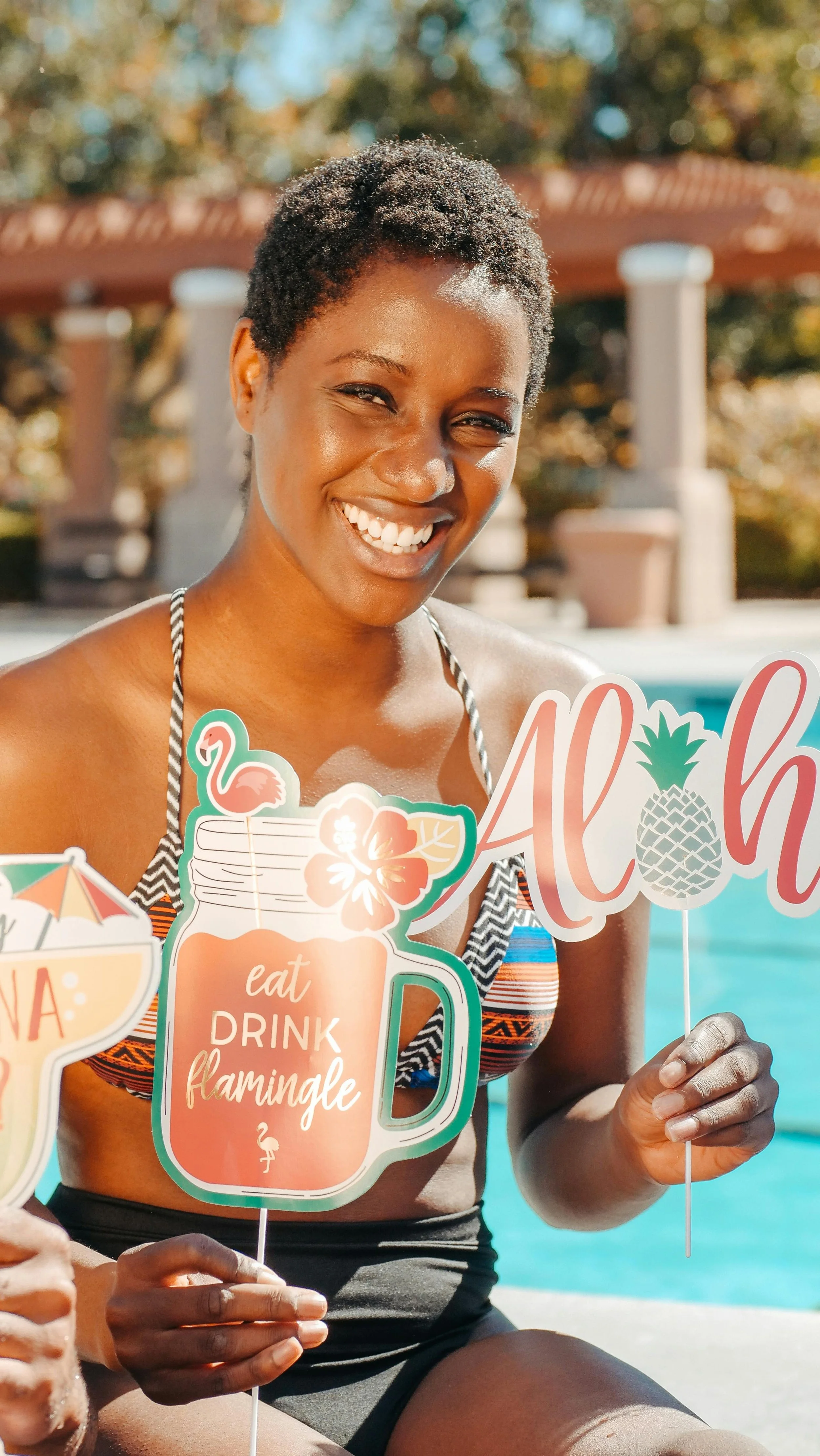 Smiling woman with short curly hair holding tropical themed photo booth props at a poolside party.